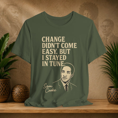 Green t-shirt with text and portrait of Sam Cooke on a wooden surface with plants and wall art in the background.