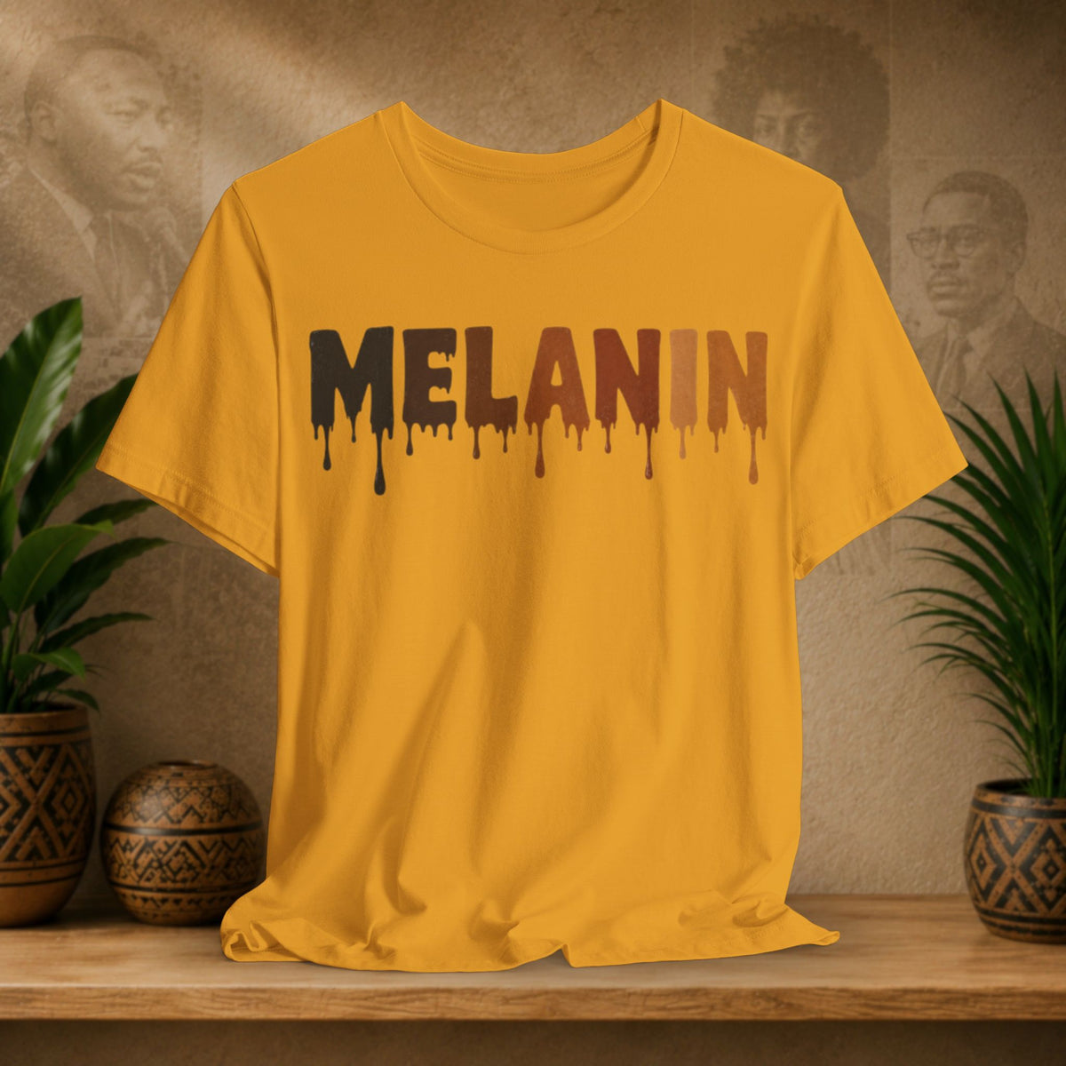A mustard yellow short-sleeve t-shirt with the word 'MELANIN' printed across the chest in a dripping paint style, styled on a wooden shelf with potted plants and a textured background.