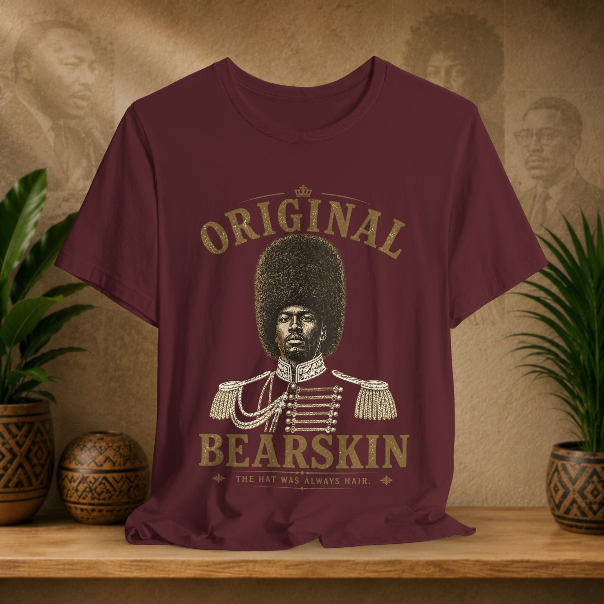 Maroon t-shirt with a graphic of a person with an afro and text 'Original Bearskin' on a wooden surface with plants and a textured wall in the background.
