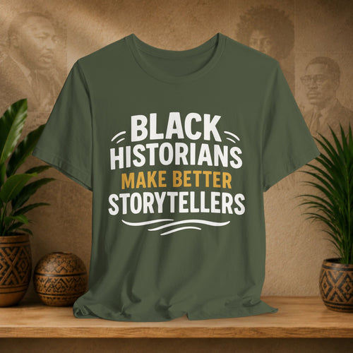 Green t-shirt with 'Black Historians Make Better Storytellers' text on a wooden surface with decorative items.