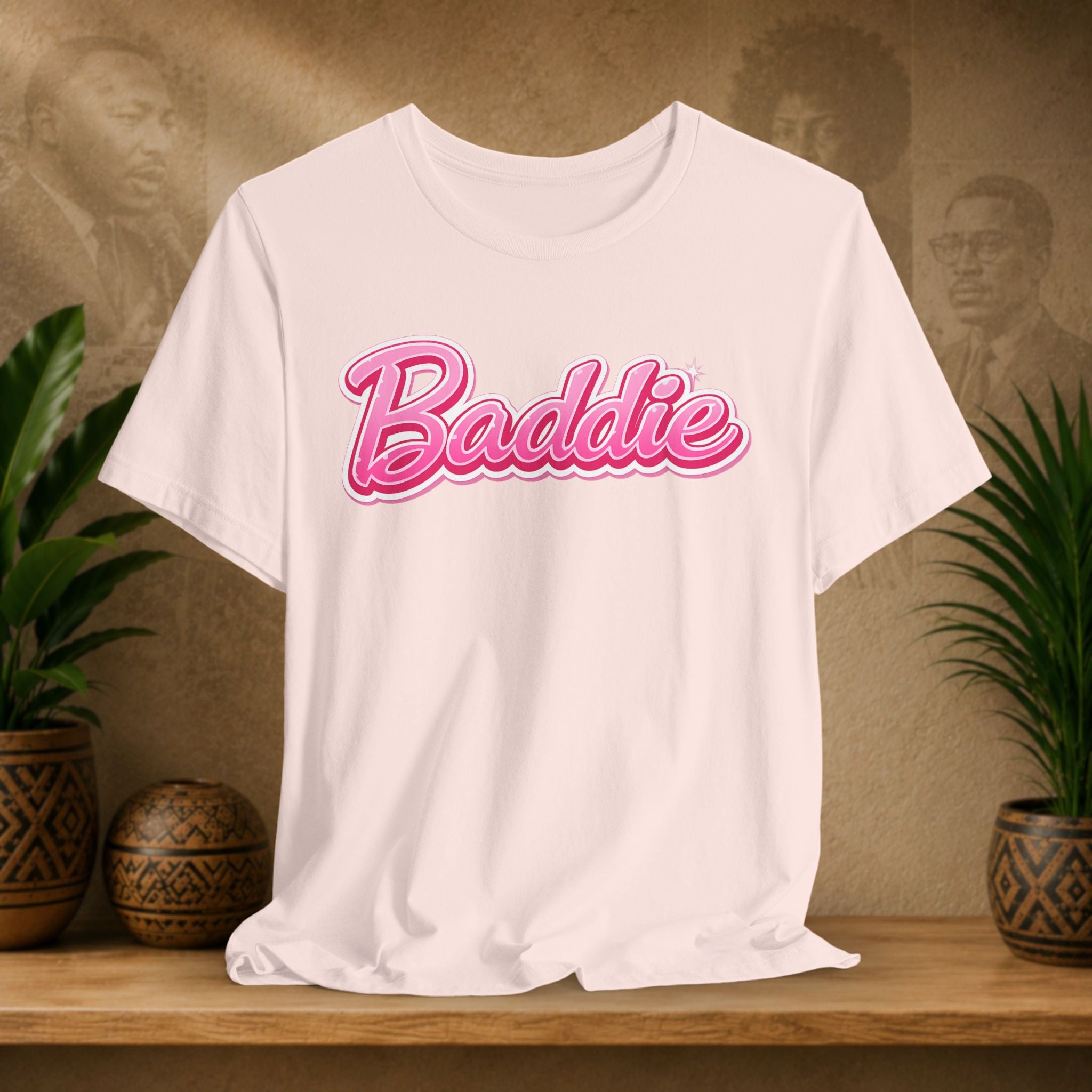 A soft pink short-sleeve T-shirt with the word 'Baddie' printed across the chest in pink cursive lettering with a white outline, displayed on a wooden shelf with potted plants and a textured background.
