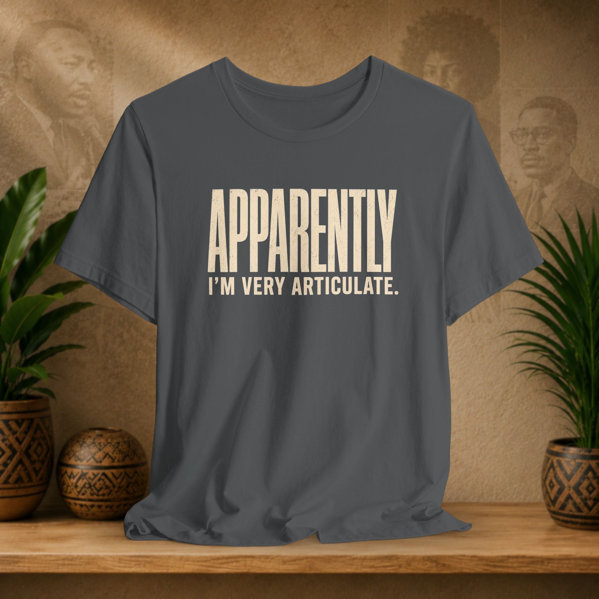 Gray t-shirt with 'APPROPRIATELY I'M VERY ARTICULATE' text on a wooden surface with plants and a textured wall background.