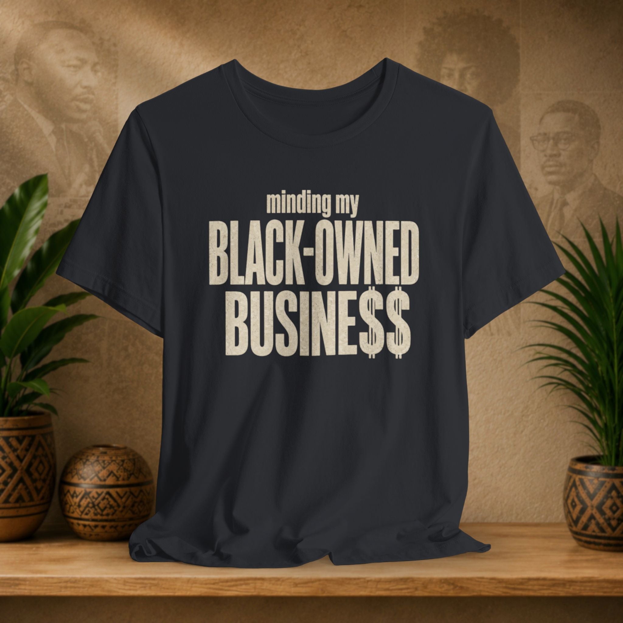 A black t-shirt with a distressed cream-colored text graphic reading 'minding my BLACK-OWNED BUSINESS$$' displayed on a wooden shelf with decorative plants and textured background.
