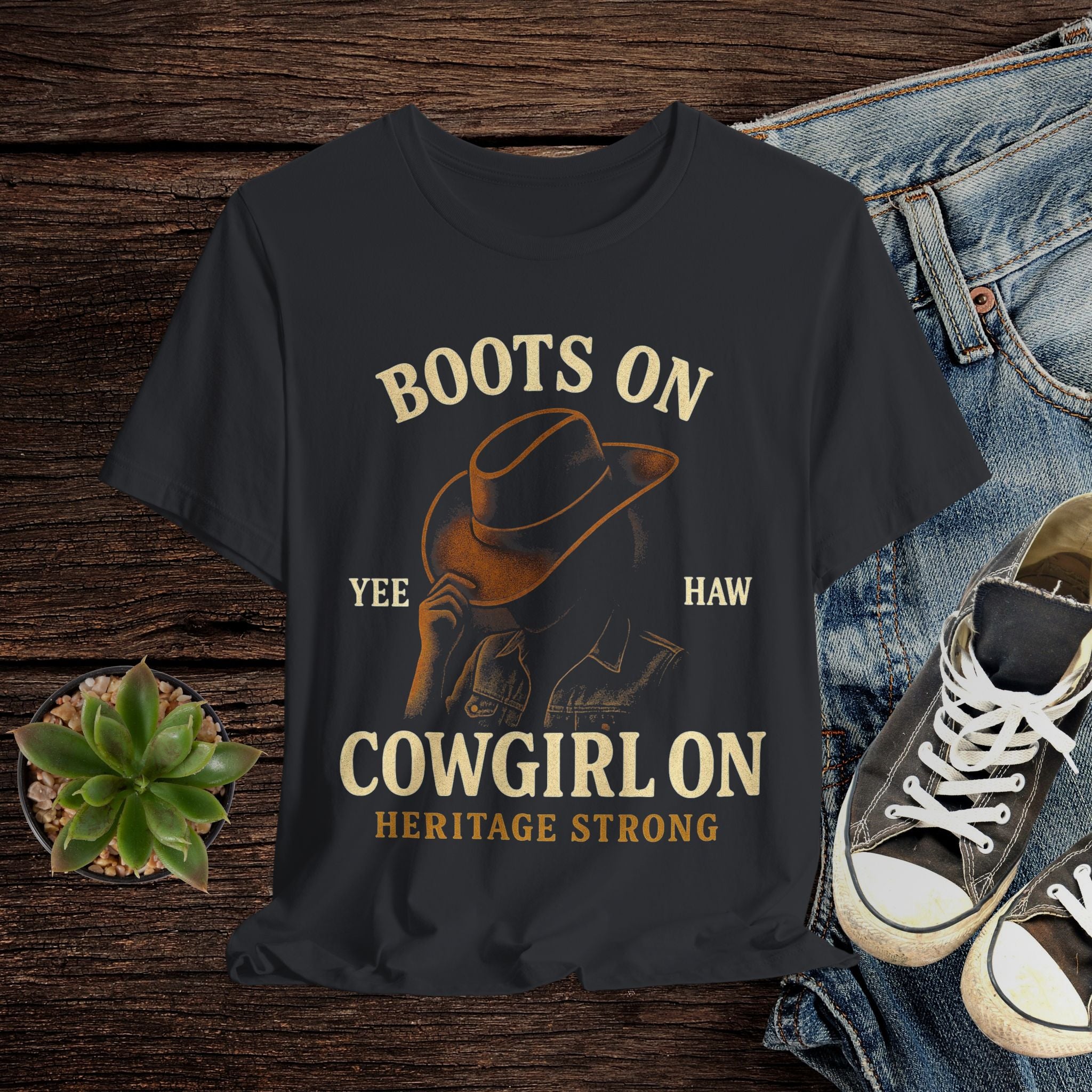 Black t-shirt with cowboy graphic and text on a wooden surface with jeans and sneakers.