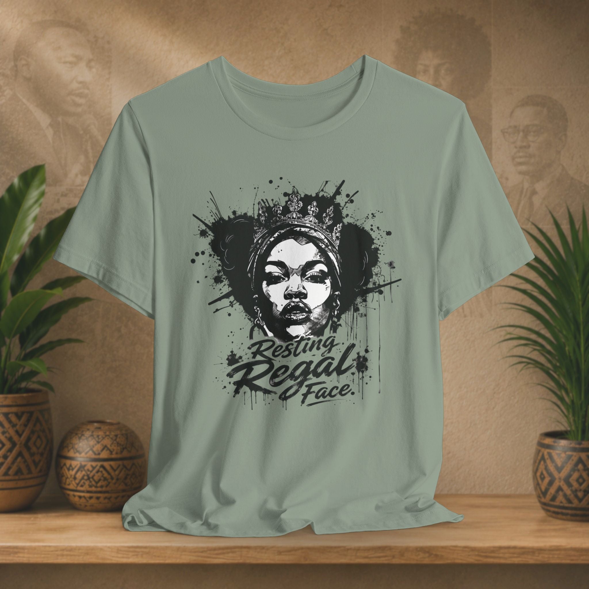 Sage green t-shirt with graphic design and text on a wooden surface with plants and decorative items.