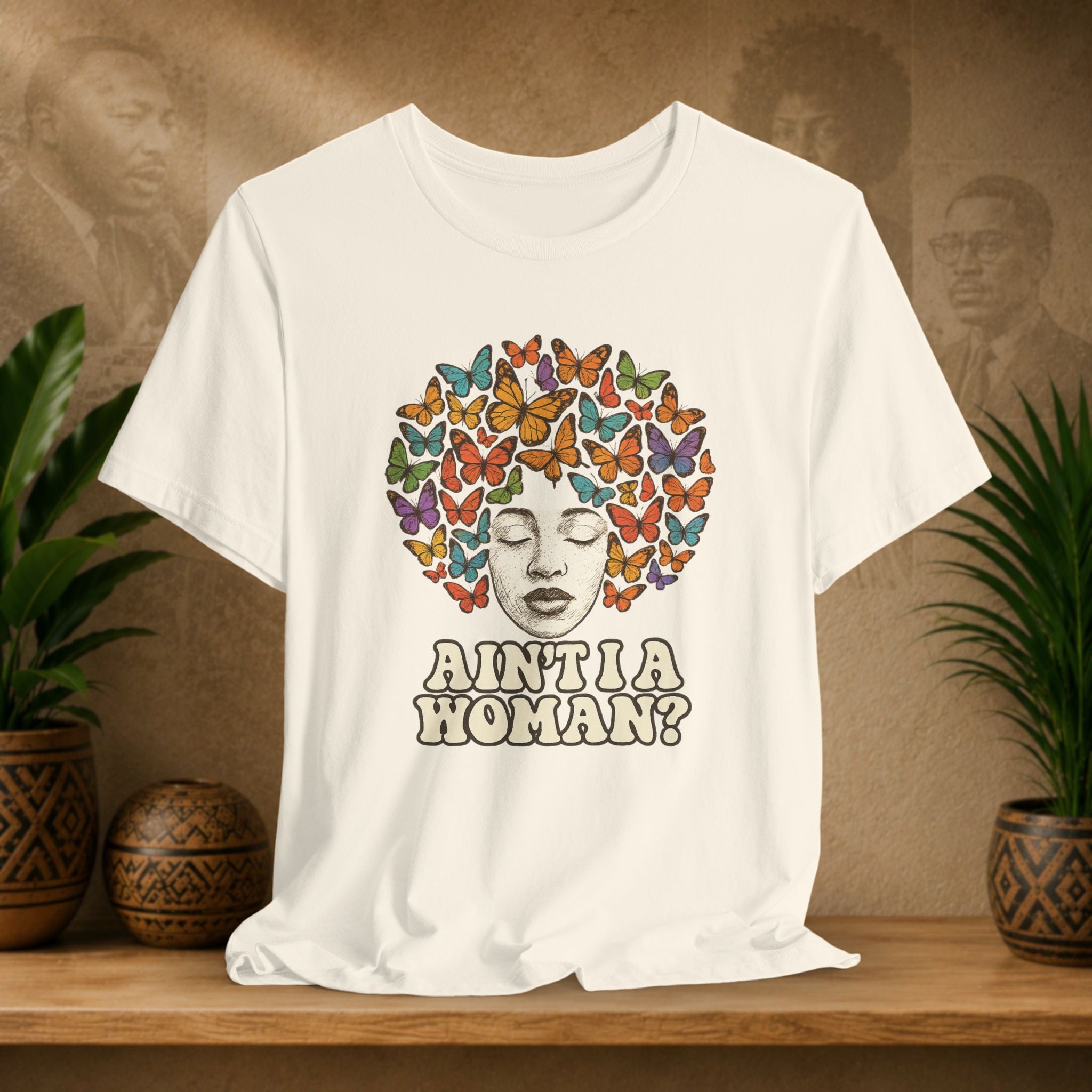 Beige t-shirt with a colorful butterfly design and text on a wooden surface with plants and a textured wall in the background.