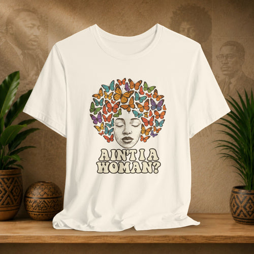 Beige t-shirt with a colorful butterfly design and text on a wooden surface with plants and a textured wall in the background.
