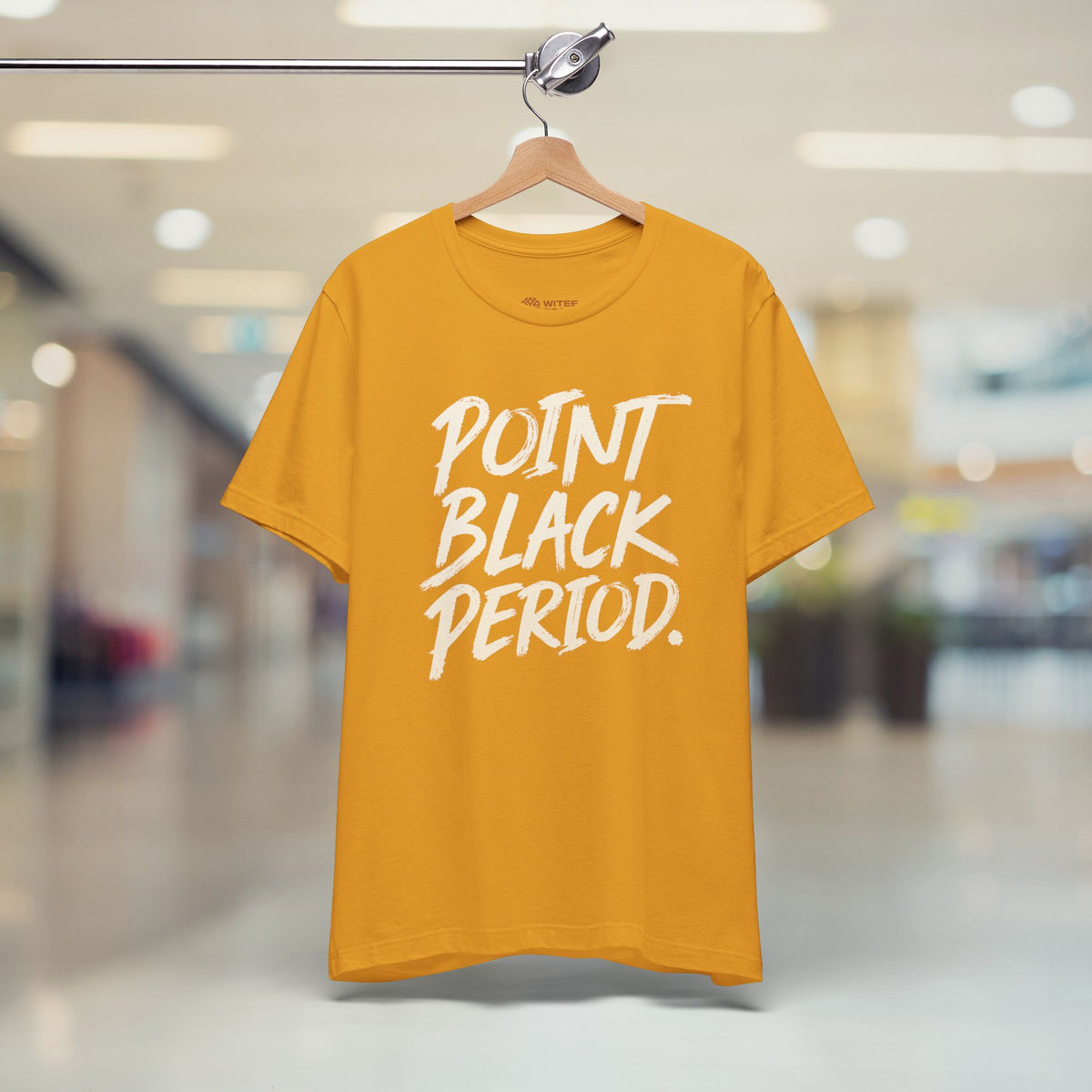 A mustard yellow t-shirt hanging on a wooden hanger from a clothing rack, featuring the text 'POINT BLACK PERIOD.' printed in white brush script across the chest.