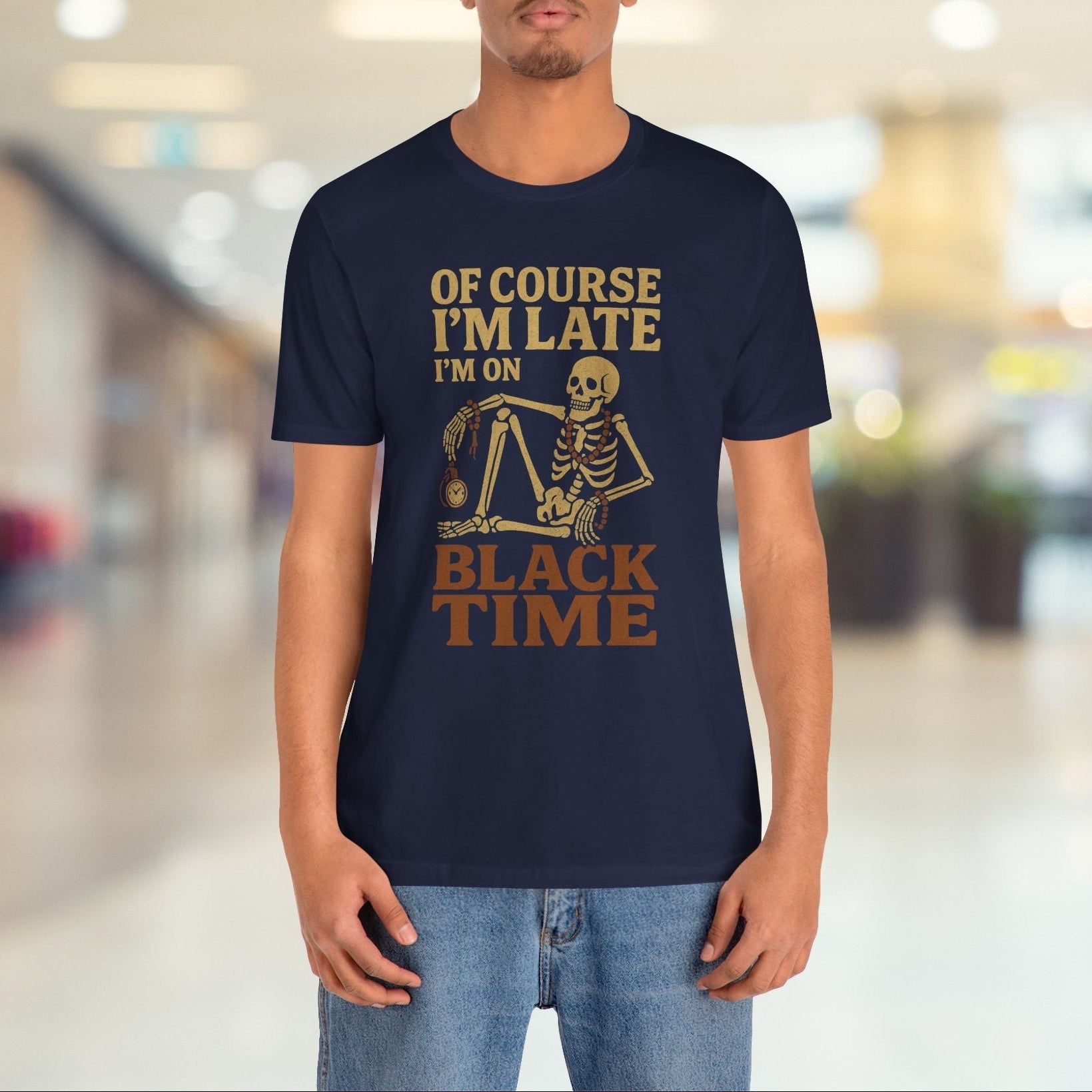 A man wearing a navy blue graphic t-shirt with a gold-colored skeleton and text reading 'OF COURSE I'M LATE I'M ON BLACK TIME', standing in a brightly lit, blurred indoor hallway.