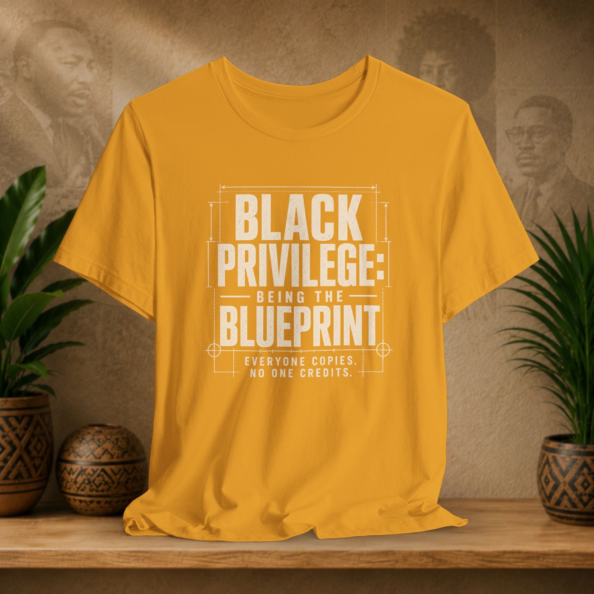 Mustard t-shirt with 'Black Privilege: Being the Blueprint' text on a wooden surface with plants and a textured wall background.