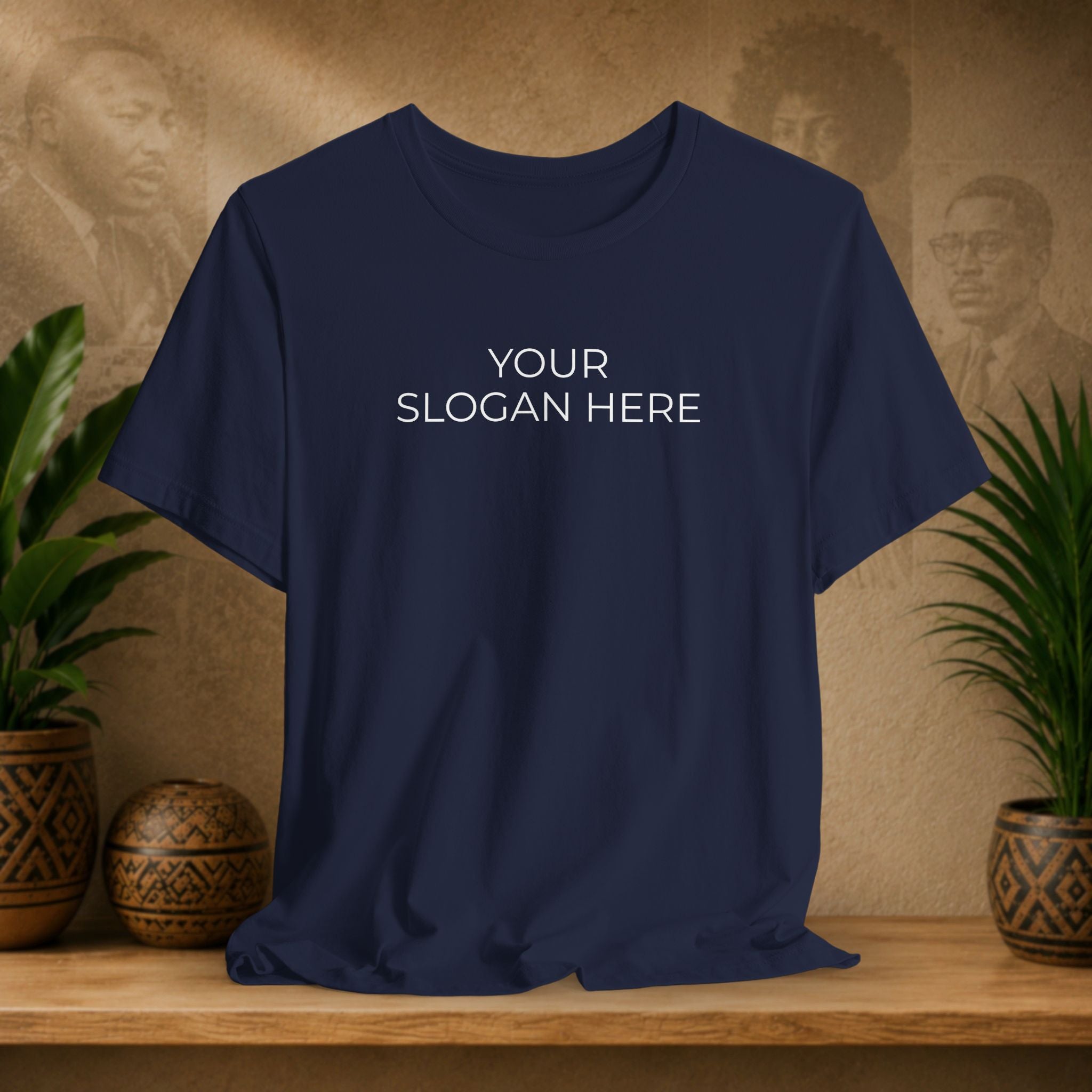A navy blue short-sleeve t-shirt with the text 'YOUR SLOGAN HERE' printed in white on the chest, displayed on a wooden shelf with decorative plants and textured background.