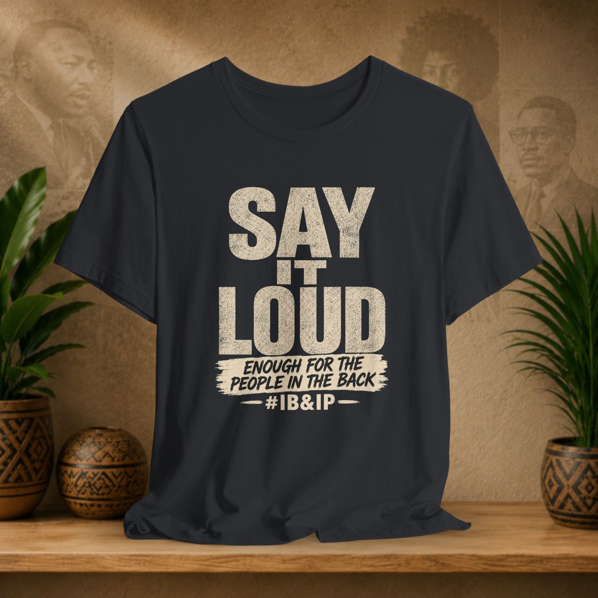 Black t-shirt with text 'Say it Loud Enough for the People in the Back #IB&IP' on a wooden surface with plants and a textured wall background.