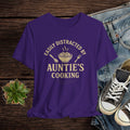 Purple t-shirt with 'Easily Distracted by Auntie's Cooking' text on a wooden surface with jeans and shoes.