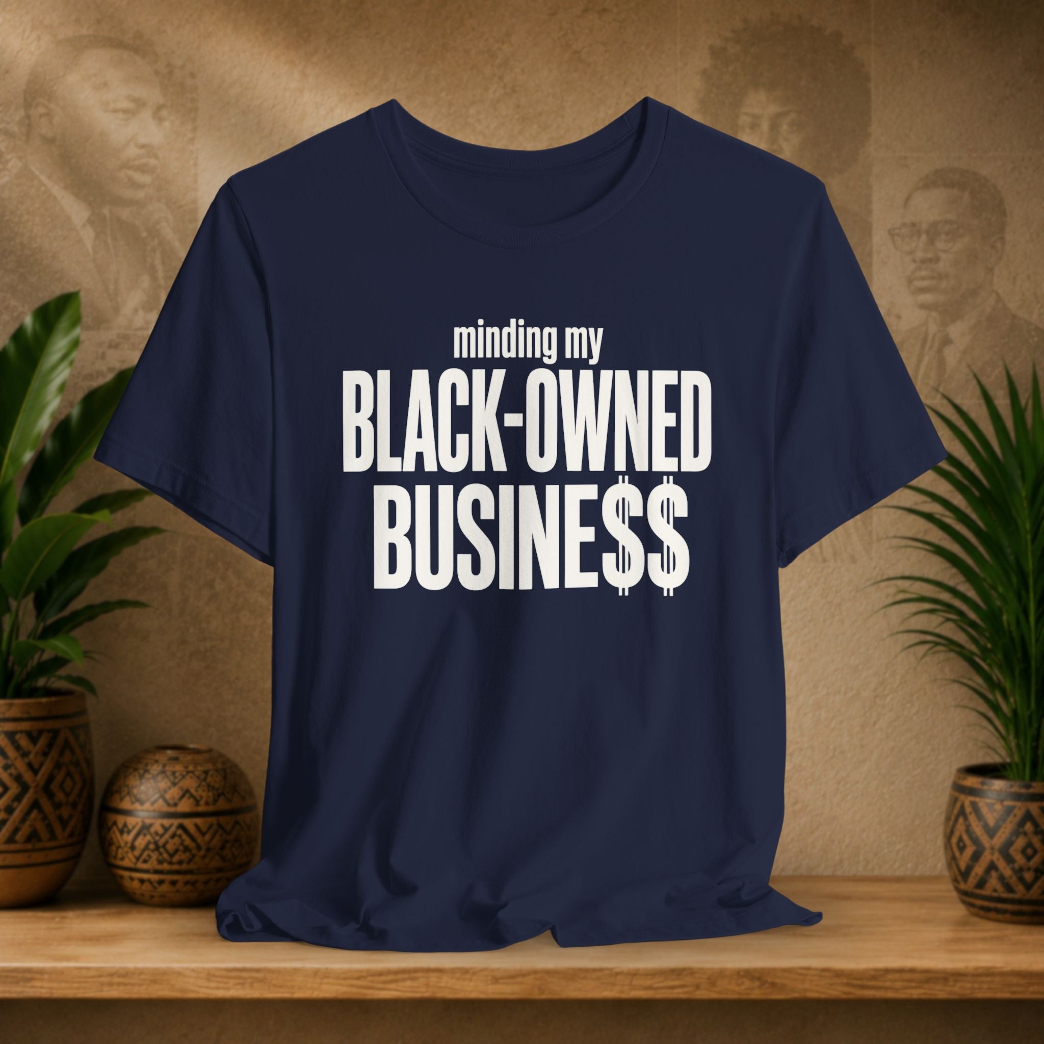 A navy blue short-sleeve t-shirt with bold white text reading 'minding my BLACK-OWNED BUSINESS$$' displayed on a wooden shelf with potted plants and a textured beige background.