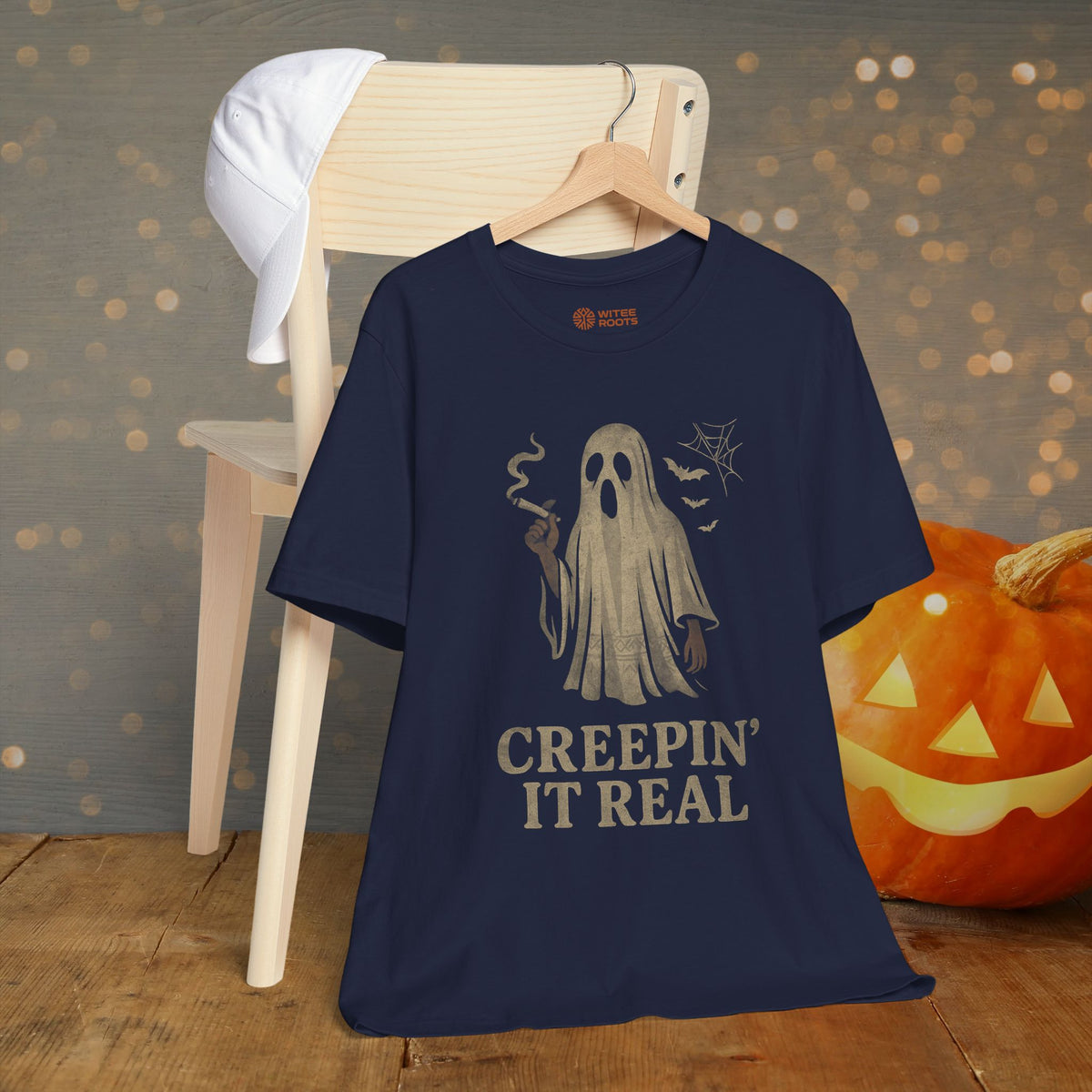 A navy blue graphic t-shirt hanging on a wooden hanger, styled in a Halloween-themed setting with a wooden chair, a white cap, and a carved pumpkin in the background.