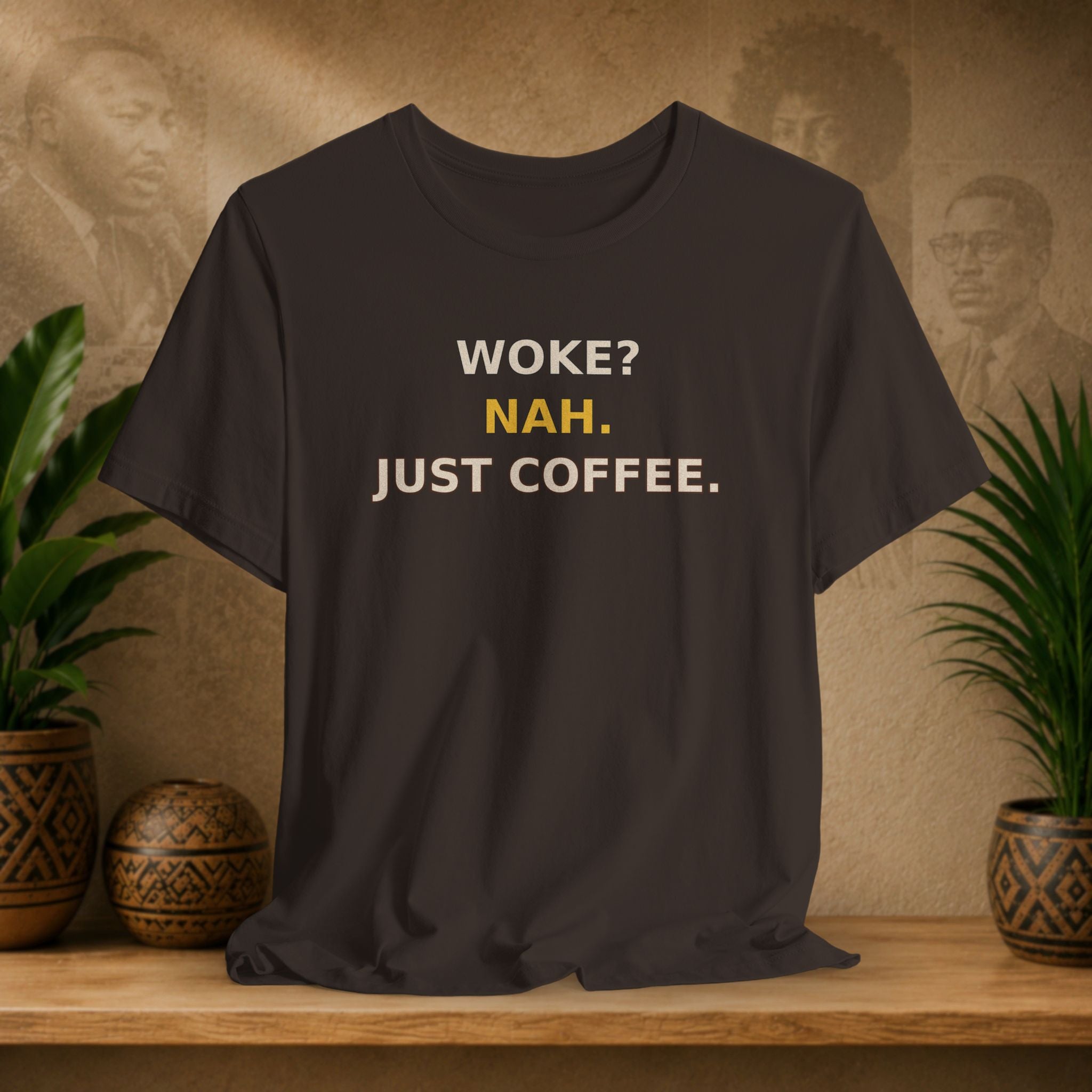 Black t-shirt with 'WOKE? NAH. JUST COFFEE.' text on a wooden surface with plants and a textured wall background.