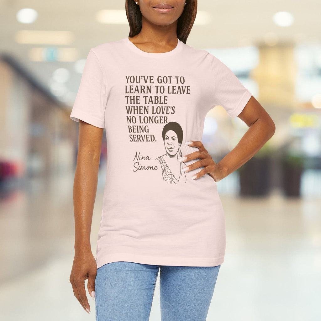 Person wearing a pink t-shirt with text and a graphic, standing in a blurred indoor setting.