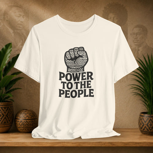 Beige t-shirt with 'Power to the People' design on a wooden surface with plants and a textured wall background.