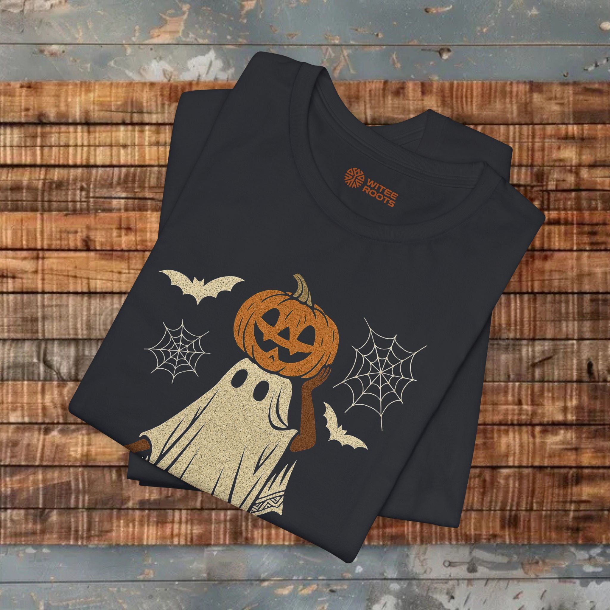 A black short-sleeve T-shirt folded on a rustic wooden surface, featuring a Halloween graphic of a jack-o'-lantern with a ghost body, white spiderwebs, and bats. The inside collar shows the brand name 'Rustle Roes'.