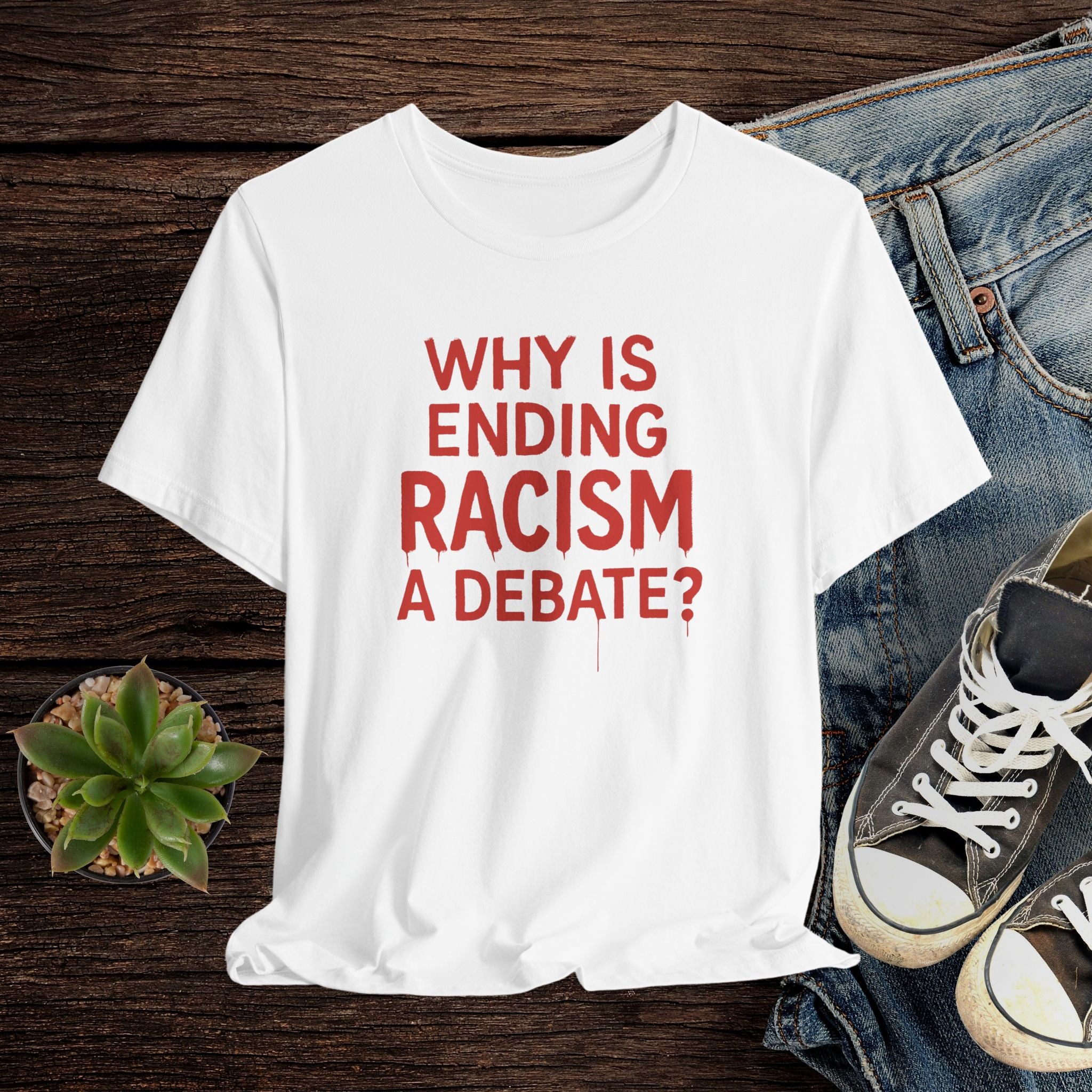 White t-shirt with red text 'WHY IS ENDING RACISM A DEBATE?' on a wooden surface with jeans and sneakers.