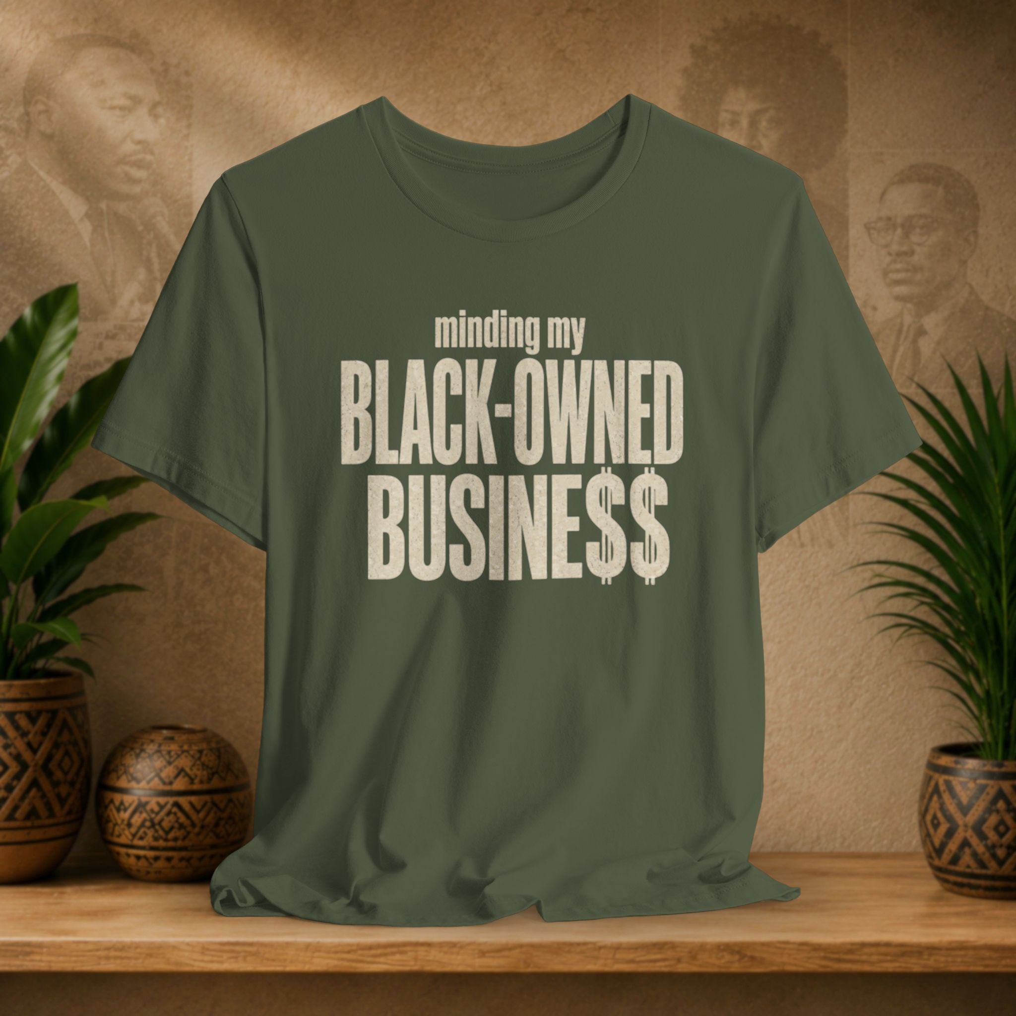 An olive green t-shirt with the text 'minding my BLACK-OWNED BUSINESS$$' printed in distressed white lettering, displayed on a wooden shelf with potted plants against a textured background.
