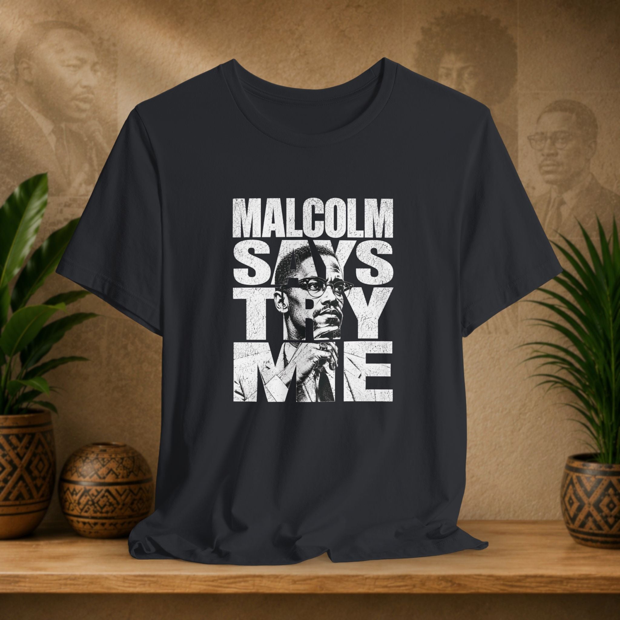 Black t-shirt with 'Malcolm Says Thy Name' graphic on a wooden surface with plants and a textured wall in the background.