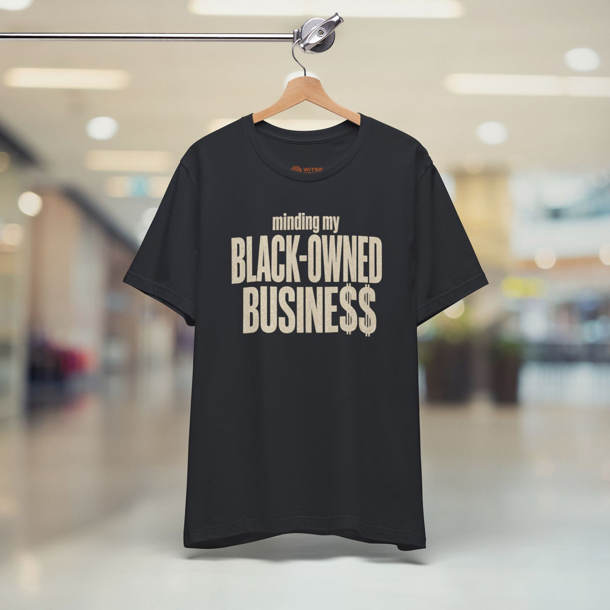 A black short-sleeve t-shirt hanging on a wooden hanger from a clothing rack, set against a blurred indoor shopping mall background. The shirt features the text 'minding my BLACK-OWNED BUSINESS$$' printed in a distressed beige font on the front.