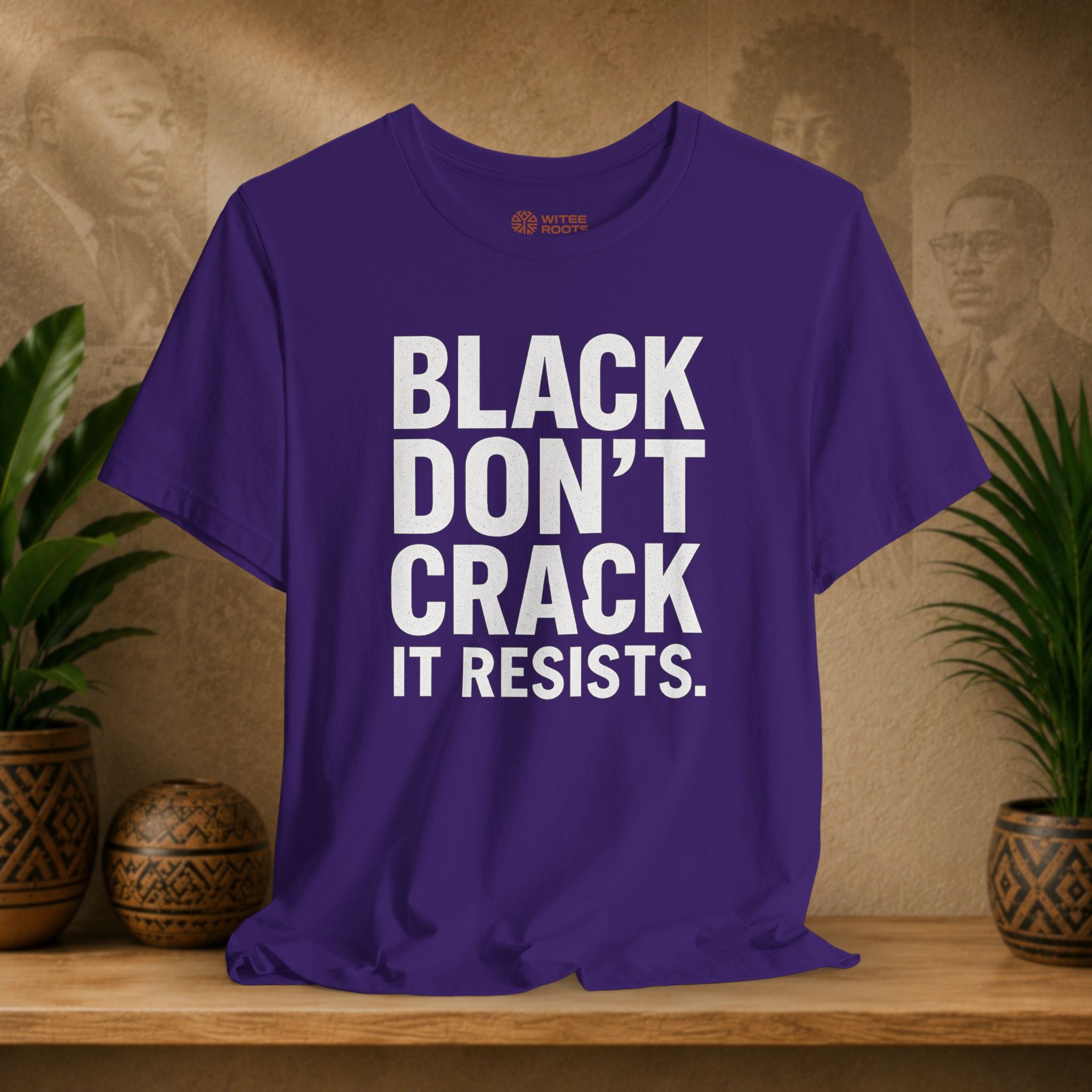 Purple t-shirt with 'BLACK DON'T CRACK IT RESISTS.' text on a wooden surface with plants and decorative items.