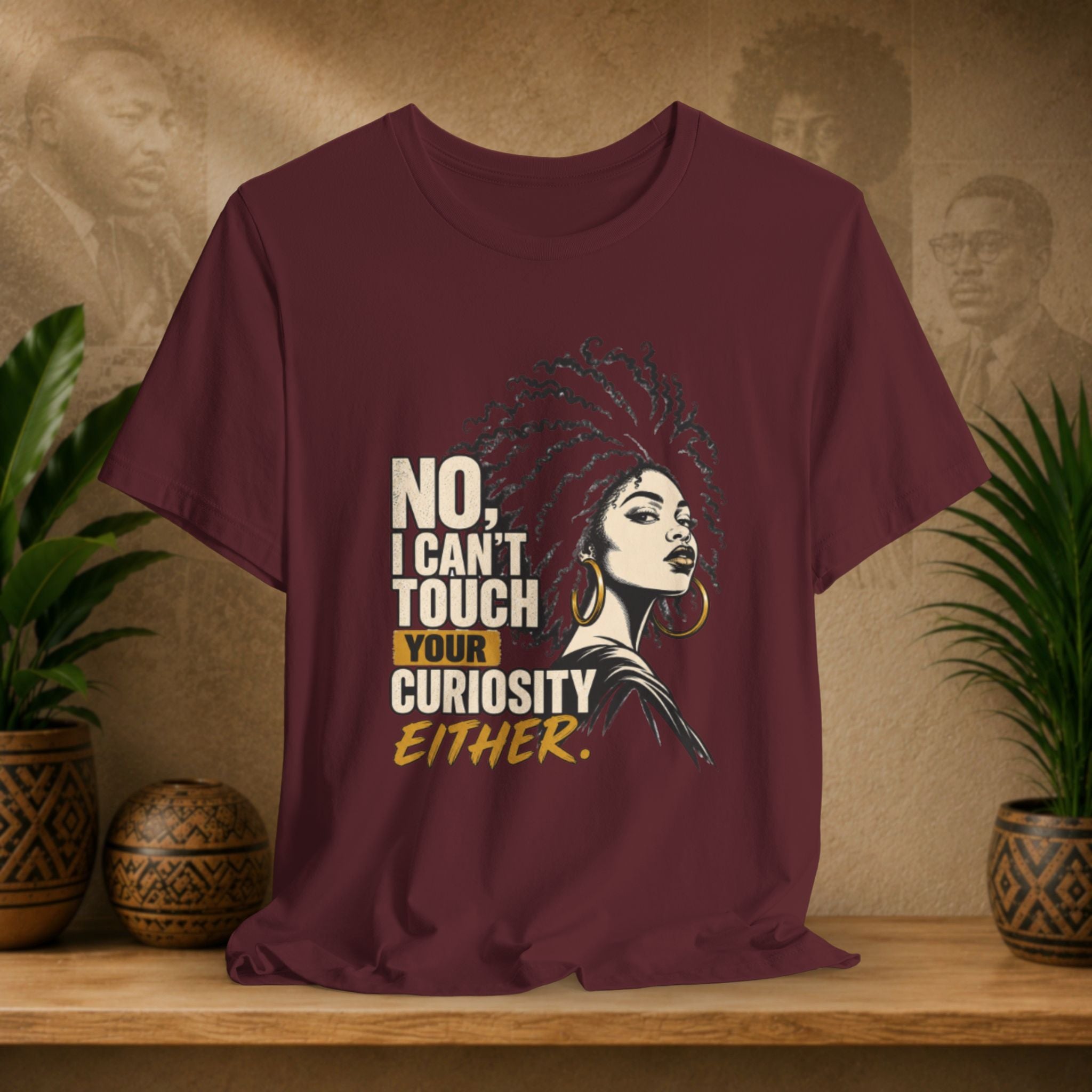 A maroon short-sleeve graphic T-shirt displayed on a wooden surface with potted plants and a textured wall behind it. The shirt features a stylized portrait of a woman with an afro and the text 'NO, I CAN'T TOUCH YOUR CURIOSITY EITHER.