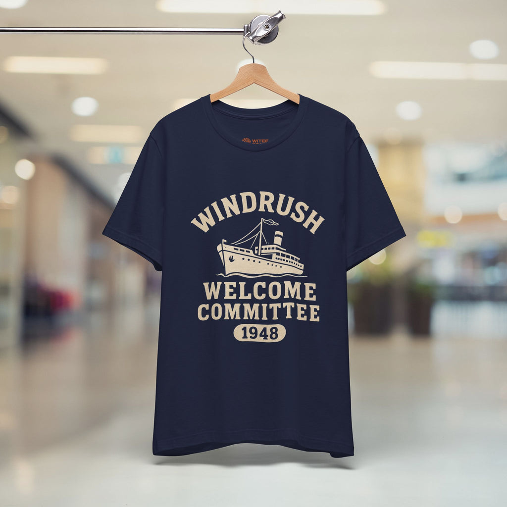 Navy blue t-shirt with 'Windrush Welcome Committee 1948' print on a hanger in a blurred indoor setting.