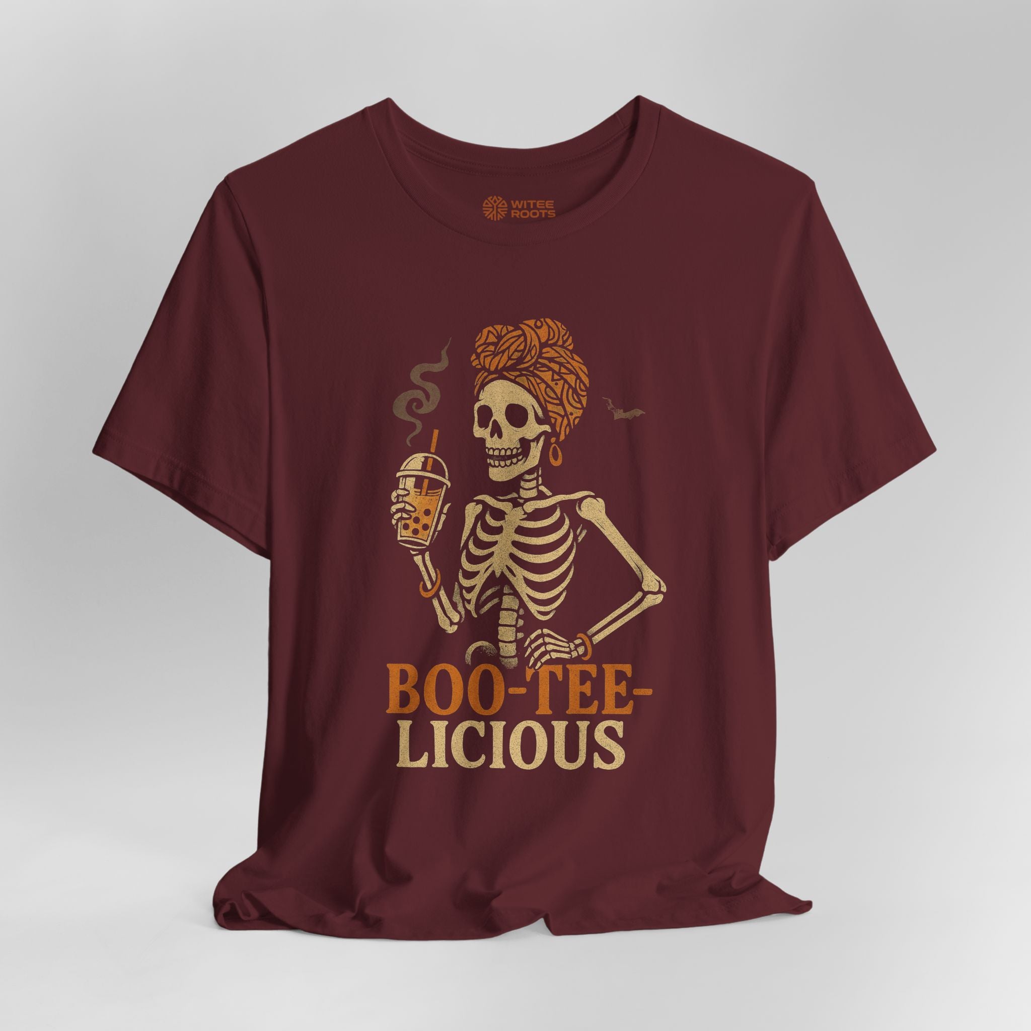 A maroon short-sleeve t-shirt with a graphic of a skeleton holding a beer mug, with the text 'BOO-TEE-LICIOUS' printed below in a vintage tan font. The shirt is displayed against a neutral gray background.