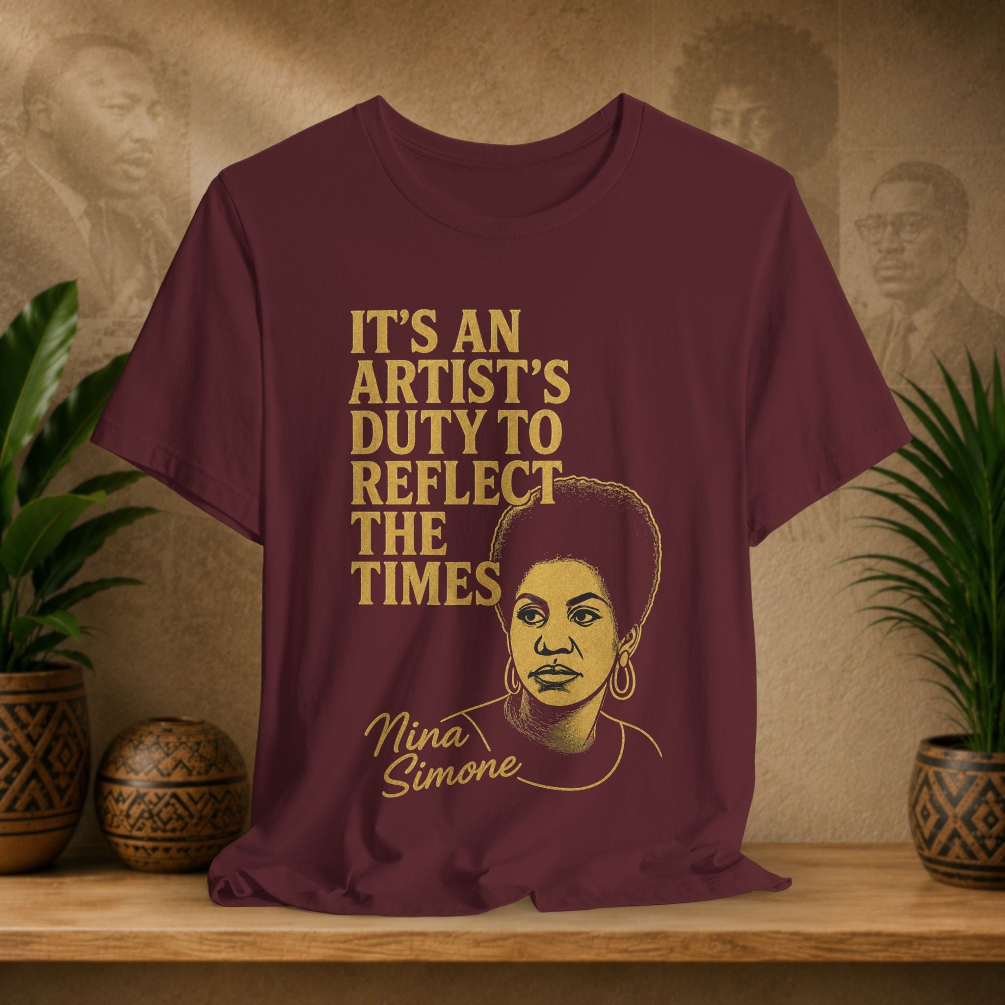 Maroon t-shirt with gold text and Nina Simone illustration on a wooden surface with plants and wall art.