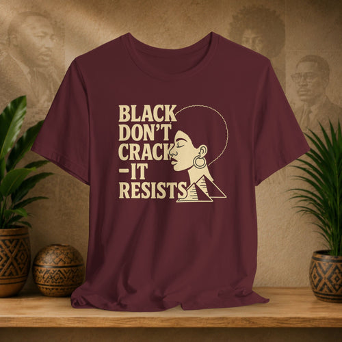 Maroon t-shirt with 'Black Don't Crack, It Resists' text and graphic on a wooden surface with decorative plants.