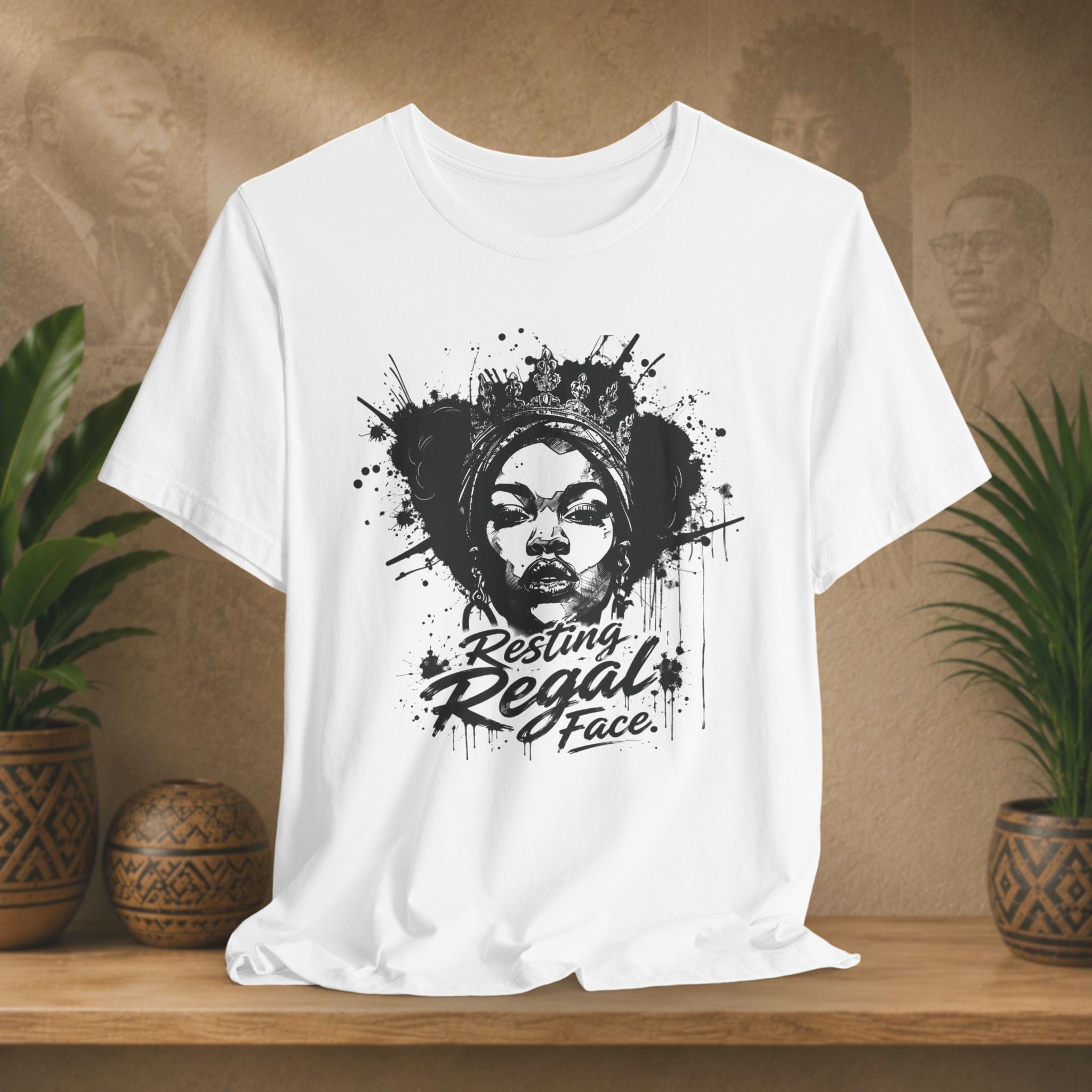 White t-shirt with a graphic design of a woman's face and text 'Resting Regal Face' on a wooden surface with plants in the background.