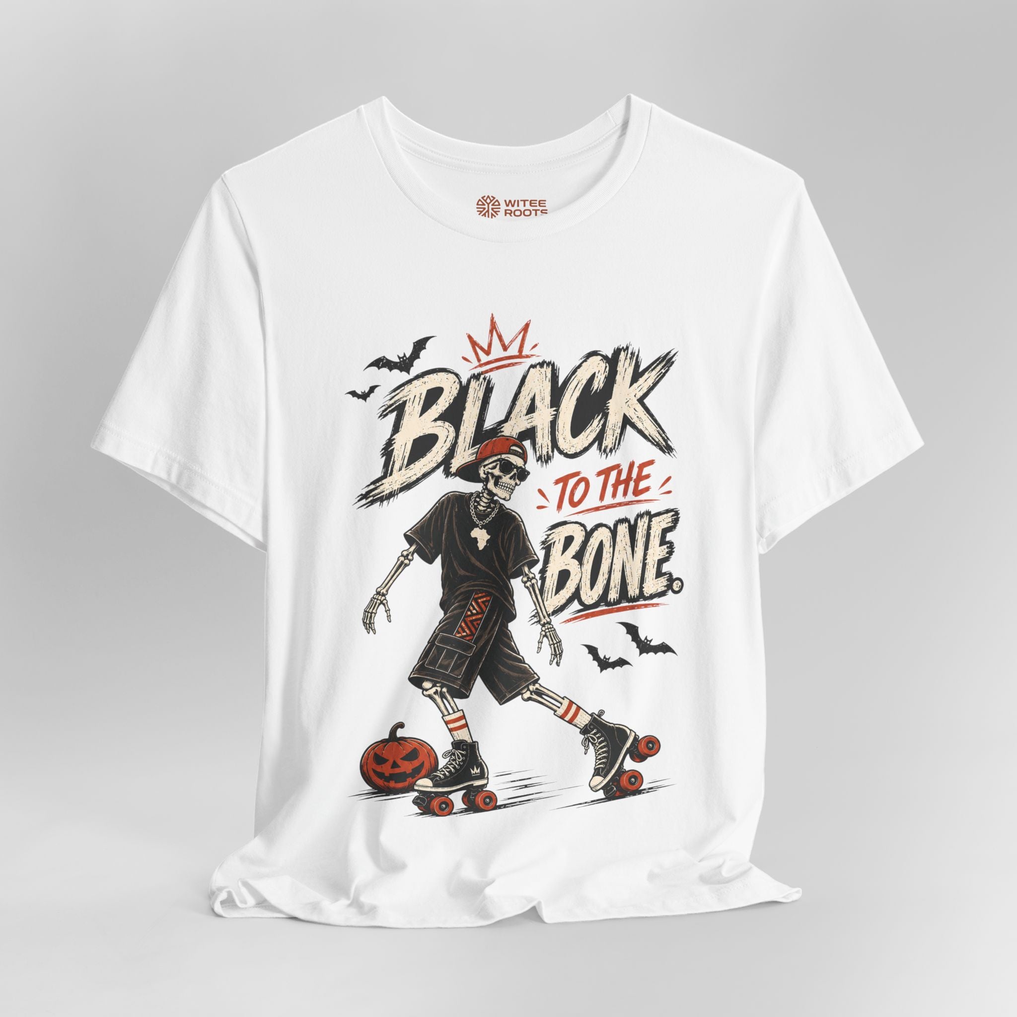 A white short-sleeve graphic T-shirt on a light gray background. The shirt features a large front print with the words 'BLACK TO THE BONE' in a distressed brush font, a crowned black-and-white skeleton roller skate, a jack-o'-lantern, and small black bats.