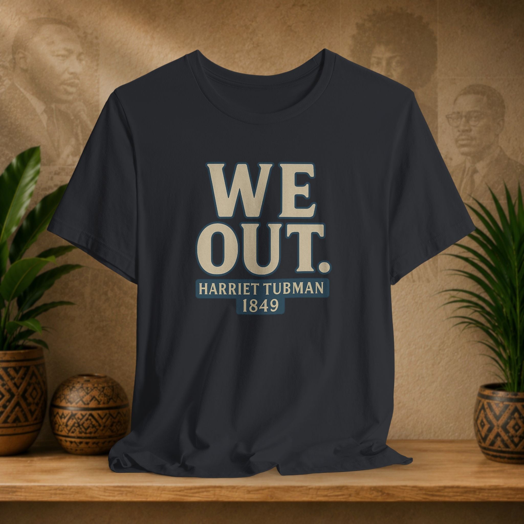 Black t-shirt with 'WE OUT. HARRETT TUBMAN 1849' text on a wooden surface with plants and a blurred background.