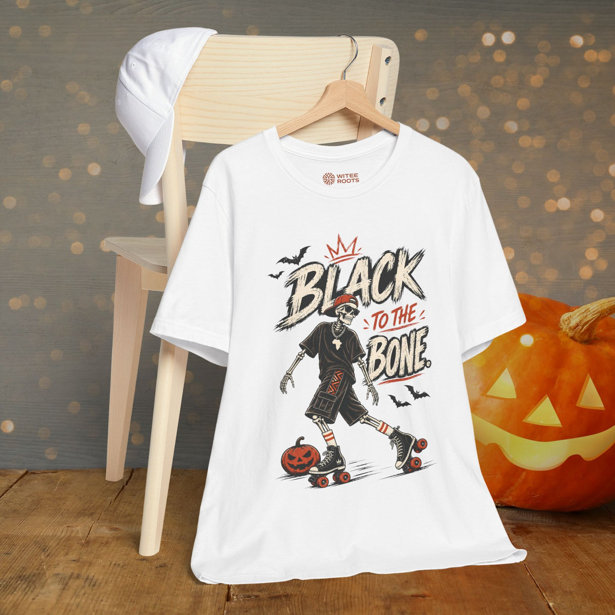 A white graphic T-shirt hanging on a wooden hanger, styled on a wooden chair with a white cap draped over the backrest. The shirt features a Halloween-themed graphic of a person roller-skating with a pumpkin, with the text 'BLACK TO THE BONE'.