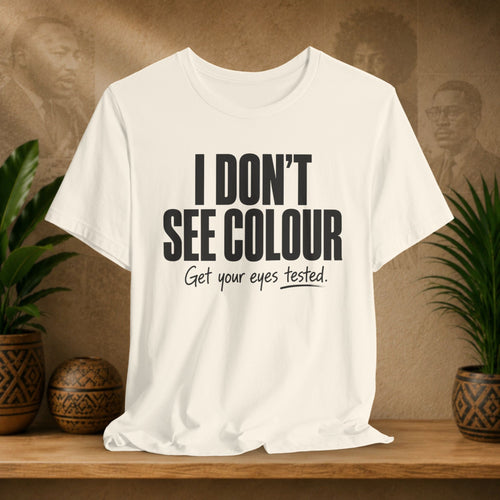 Beige t-shirt with text 'I DON'T SEE COLOUR Get your eyes tested.' on a wooden surface with plants and a blurred background.
