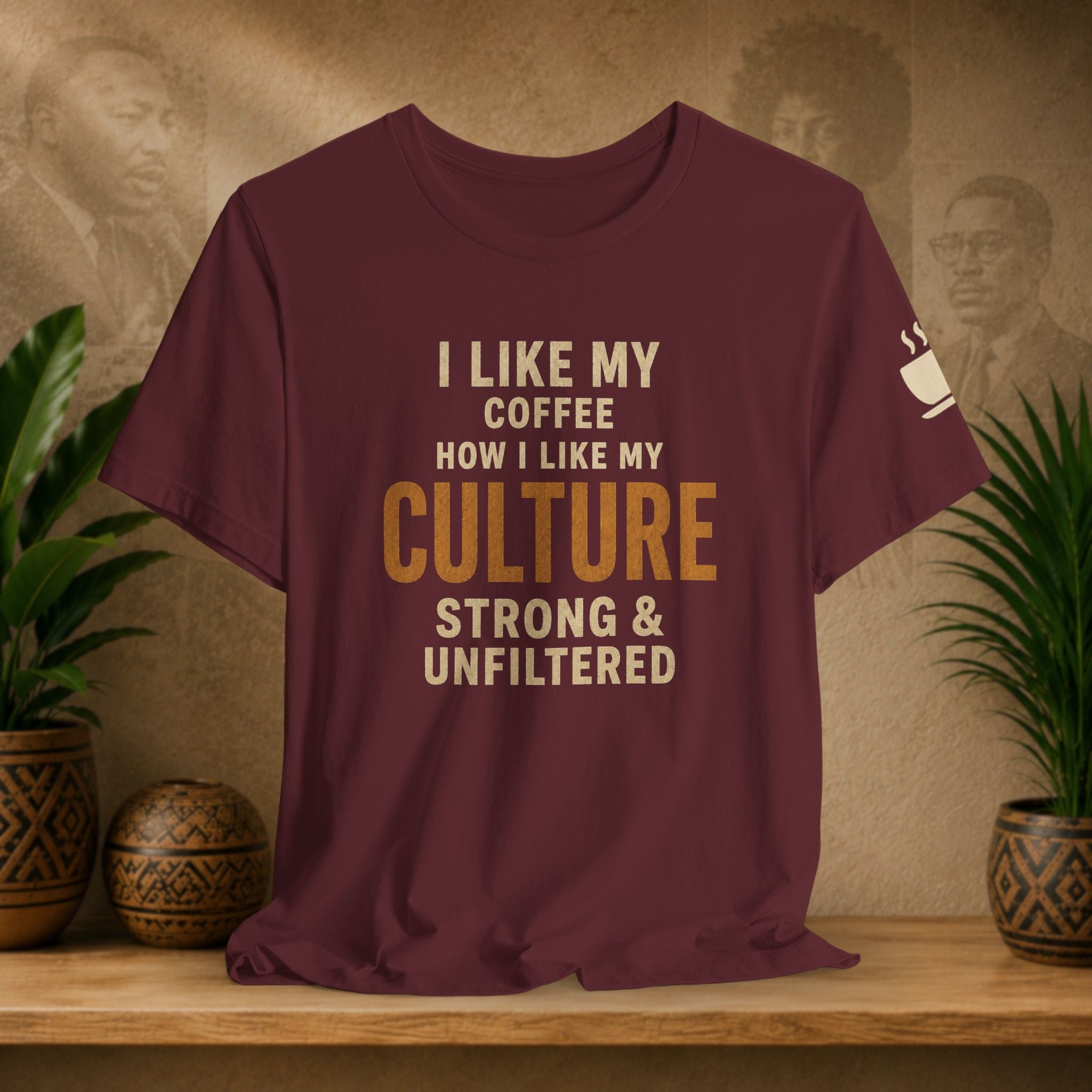 A maroon short-sleeve t-shirt with a humorous coffee-themed graphic reading 'I LIKE MY COFFEE HOW I LIKE MY CULTURE STRONG & UNFILTERED' in cream and gold lettering, displayed on a wooden shelf with potted plants.