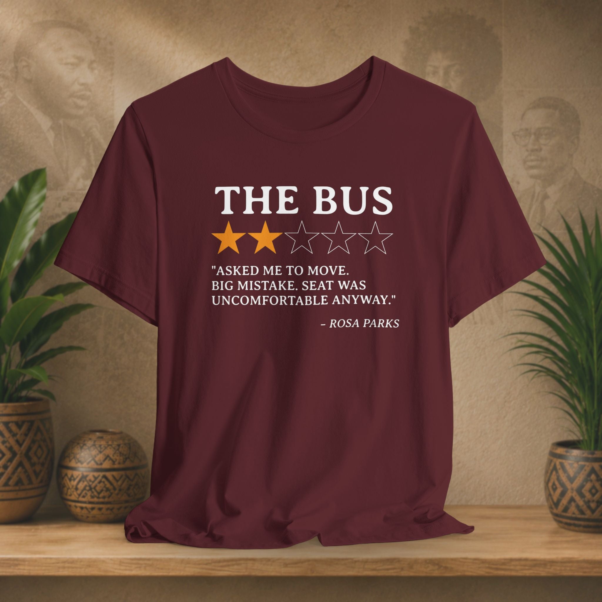 Maroon  t-shirt with a quote on it displayed on a wooden surface with plants and a wall in the background.