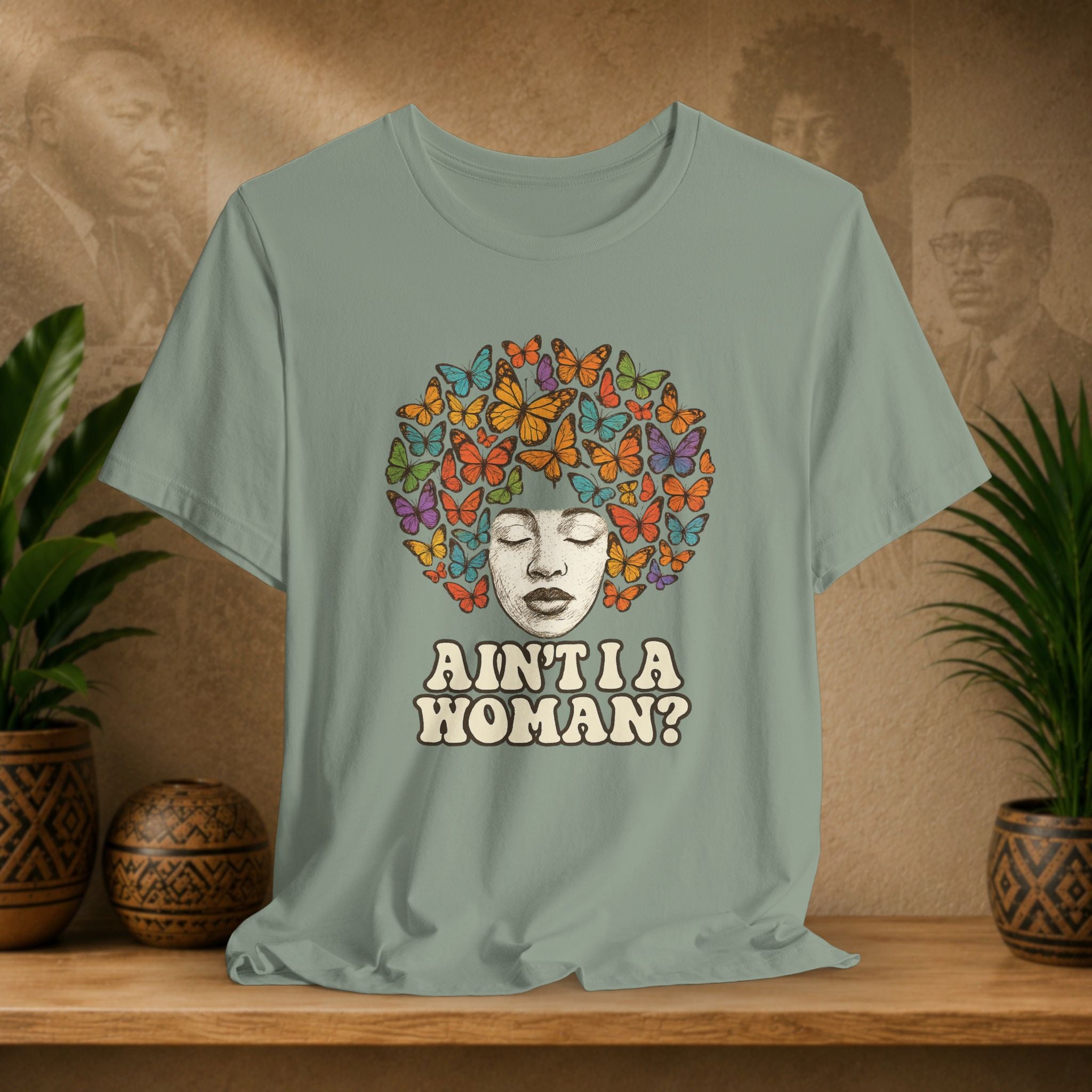 A sage green graphic t-shirt displayed on a wooden shelf. The shirt features a retro-style illustration of a Black woman's face surrounded by a vibrant, multicolored afro made of butterflies, with the text 'AIN'T I A WOMAN?' printed below in a beige retro font.