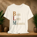 A beige short-sleeve graphic T-shirt displayed on a wooden surface, featuring the text 'BLACK LOVED MELANATED' in stacked, stylized letters. The background is a warm brown wall with faint portraits, flanked by potted green plants.