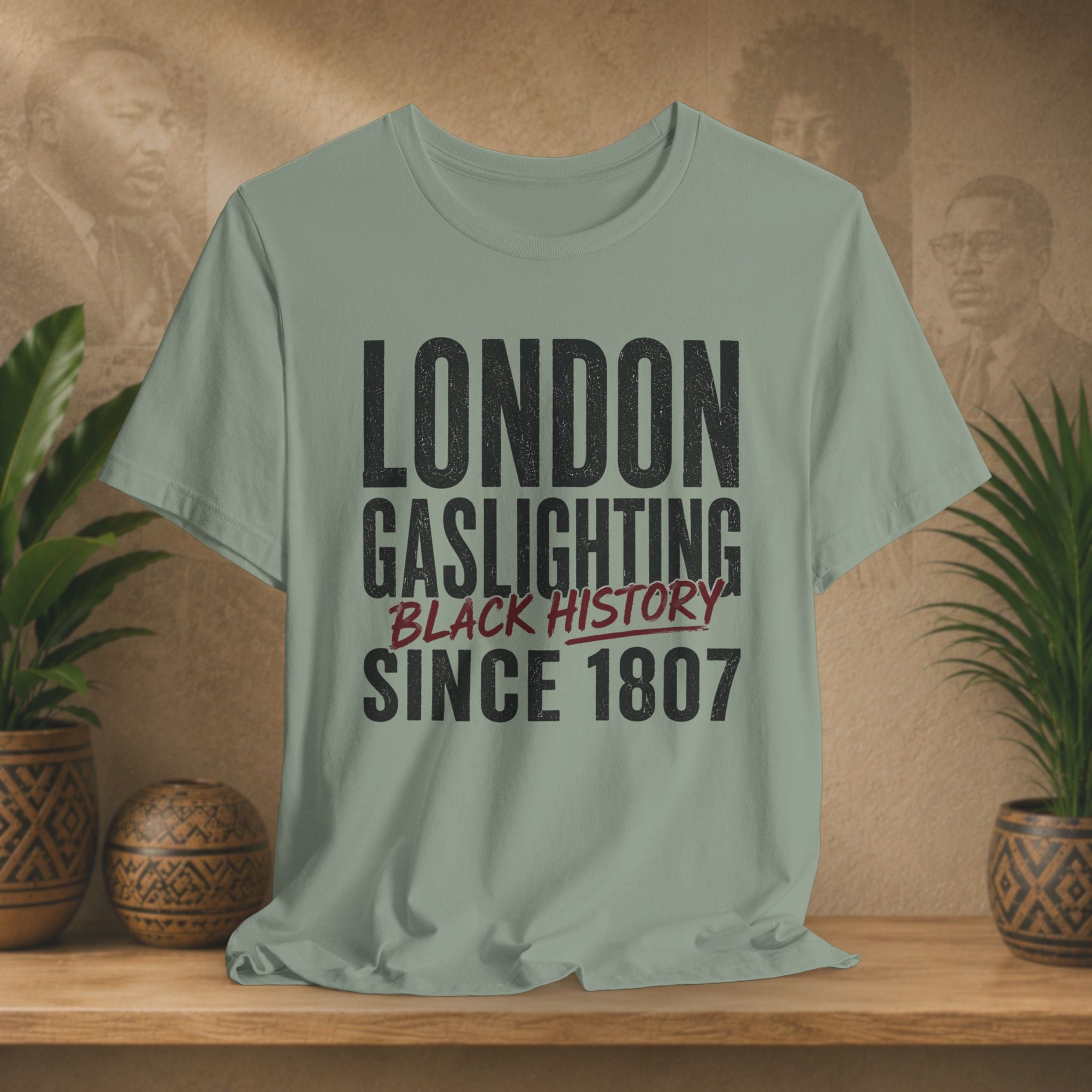 Sage t-shirt with 'London Gaslighting Black History Since 1807' text on a wooden surface with plants and a blurred background.