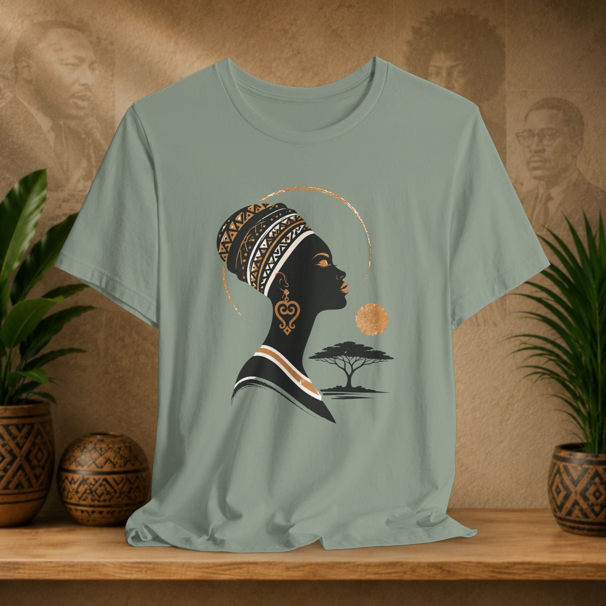 Sage green t-shirt with a graphic of a woman's silhouette, tree, and sun on a wooden surface with plants and decorative items.