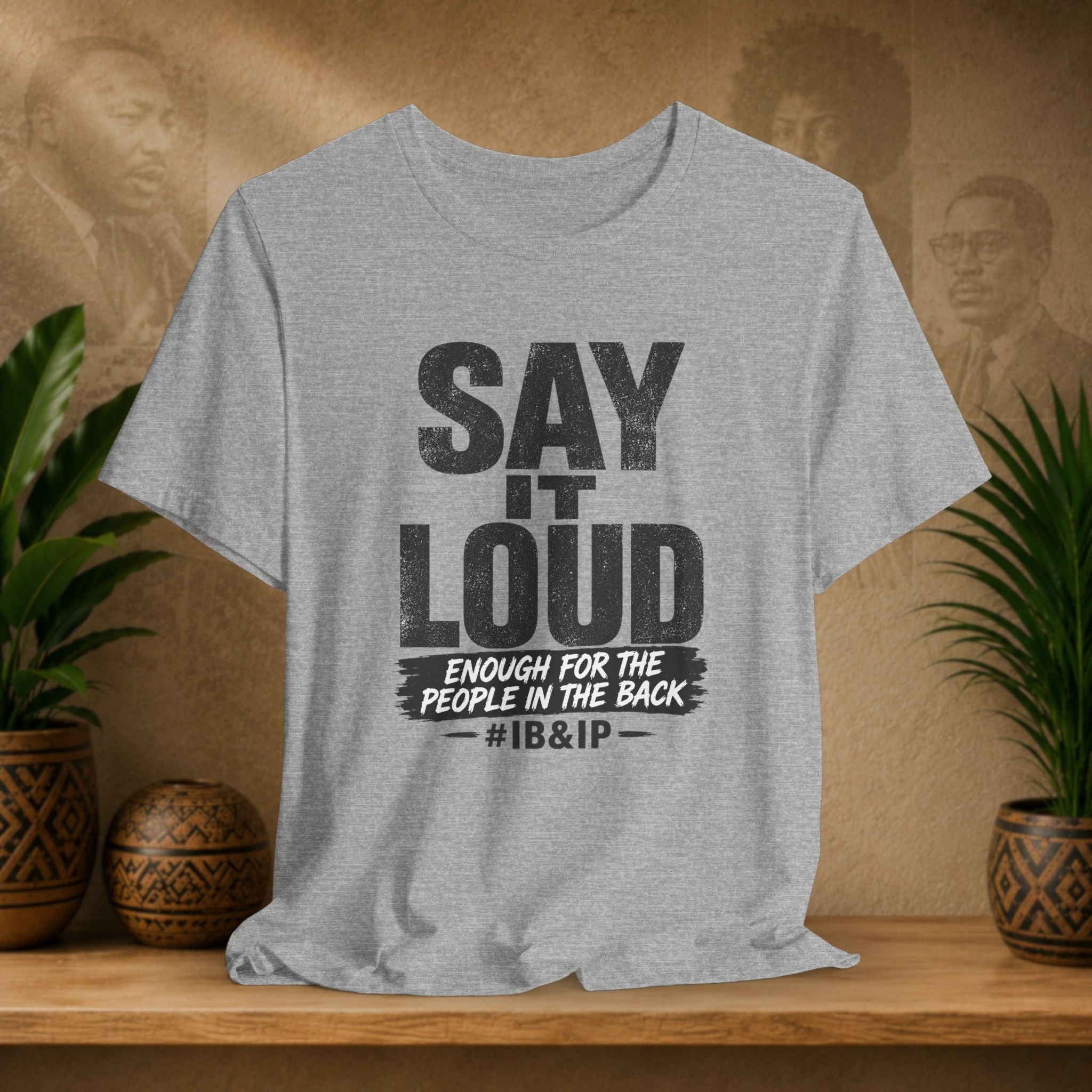 Grey t-shirt with text 'Say it Loud Enough for the People in the Back #IB&IP' on a wooden surface with plants and a textured wall background.