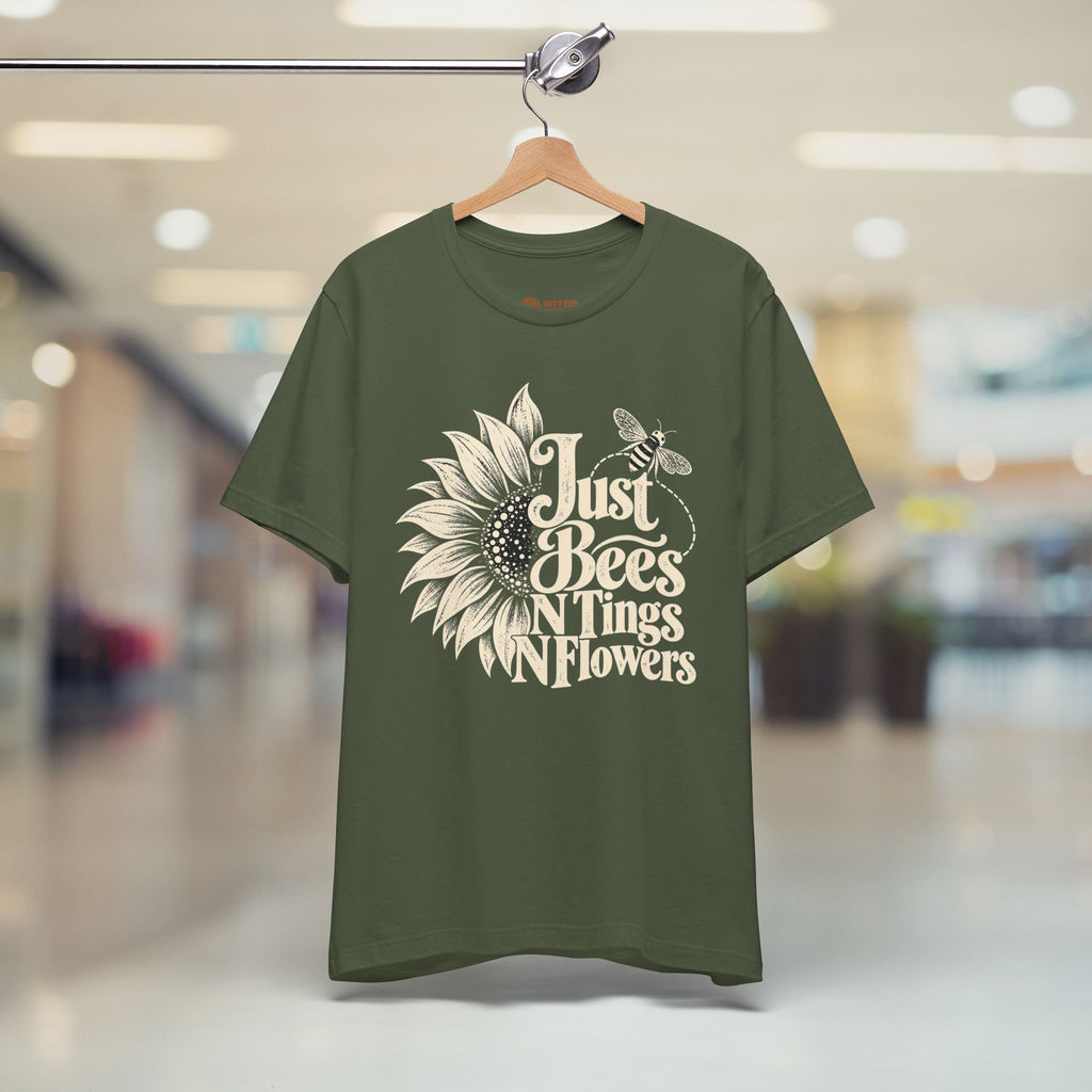 An olive green short-sleeve t-shirt hanging on a wooden hanger from a clothing rack, featuring a cream-colored graphic of a sunflower, bee, and the text 'Just Bees N Tings N Flowers' against a blurred indoor shopping mall background.
