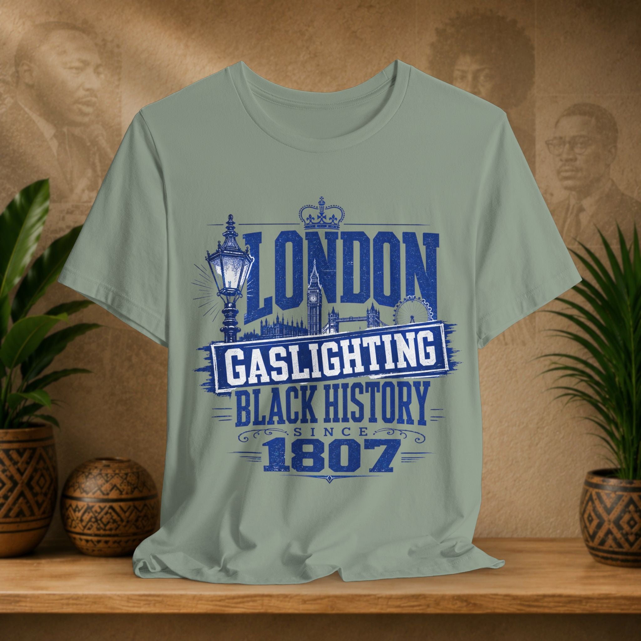 Sage t-shirt with 'London Gaslighting Black History Since 1807' design on a wooden surface with plants and a blurred background.