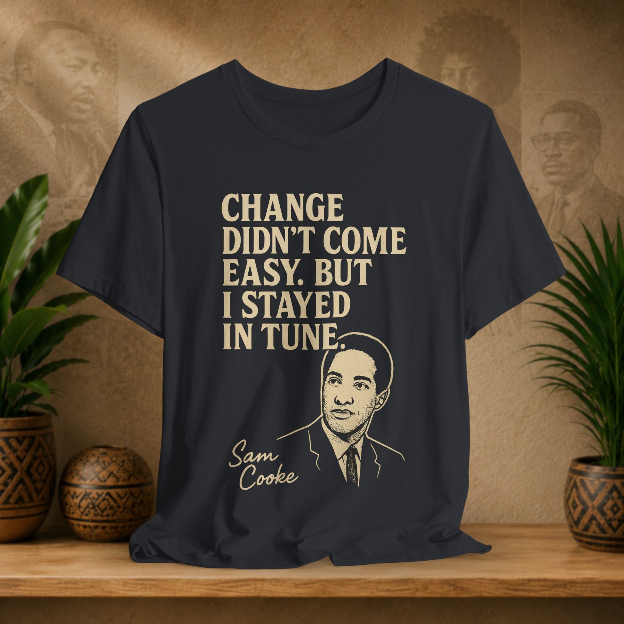 Black t-shirt with text and a portrait on a wooden surface with plants and a wall with portraits in the background