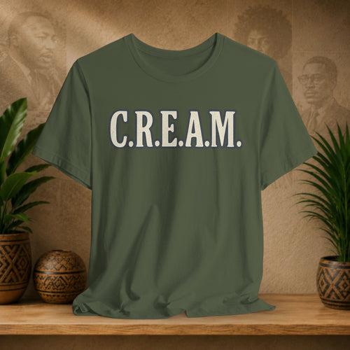 Green t-shirt with 'C.R.E.A.M.' text on a wooden surface with decorative plants and background.
