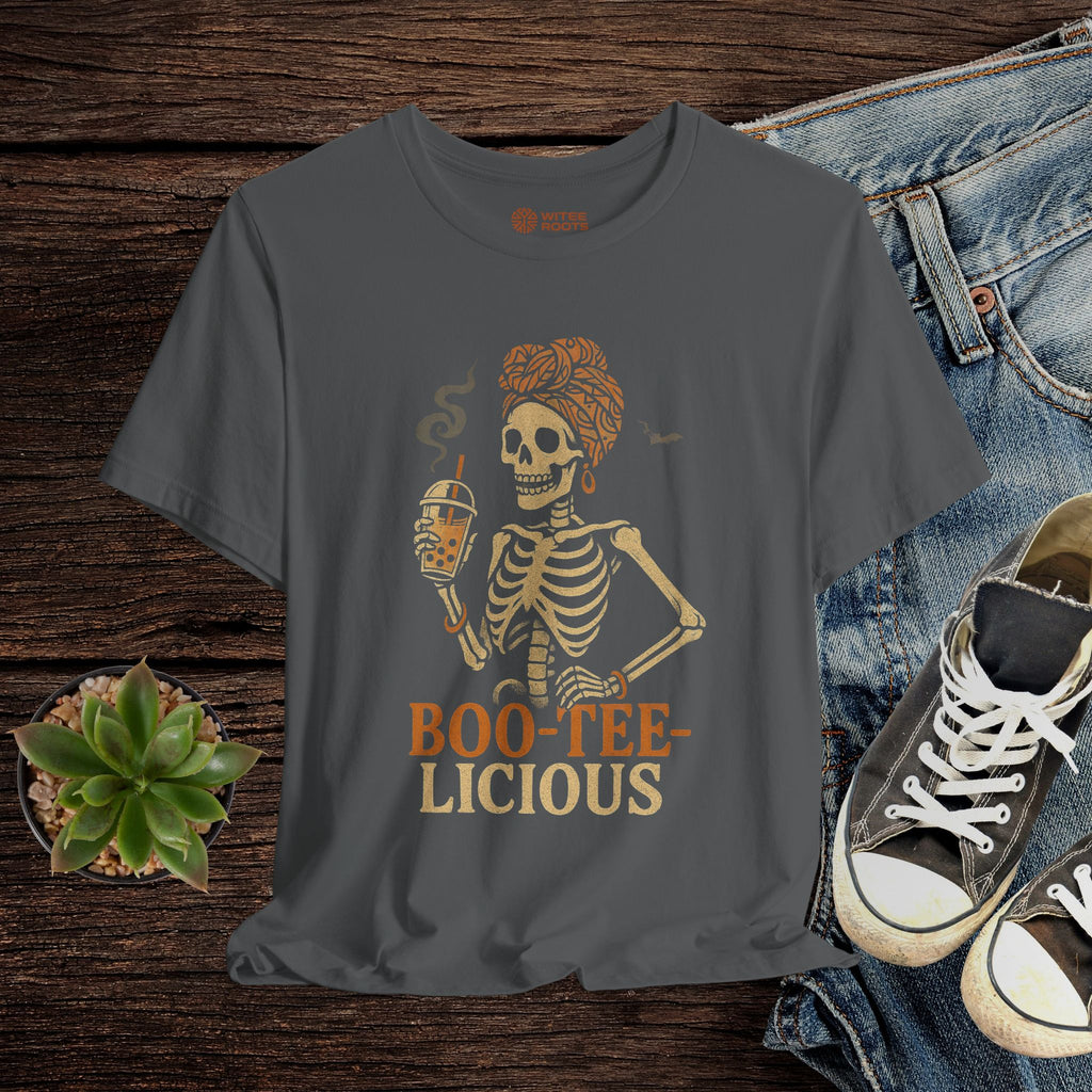 Gray t-shirt with skeleton design and 'BOO-TEE-LICIOUS' text on a wooden surface with jeans and sneakers.